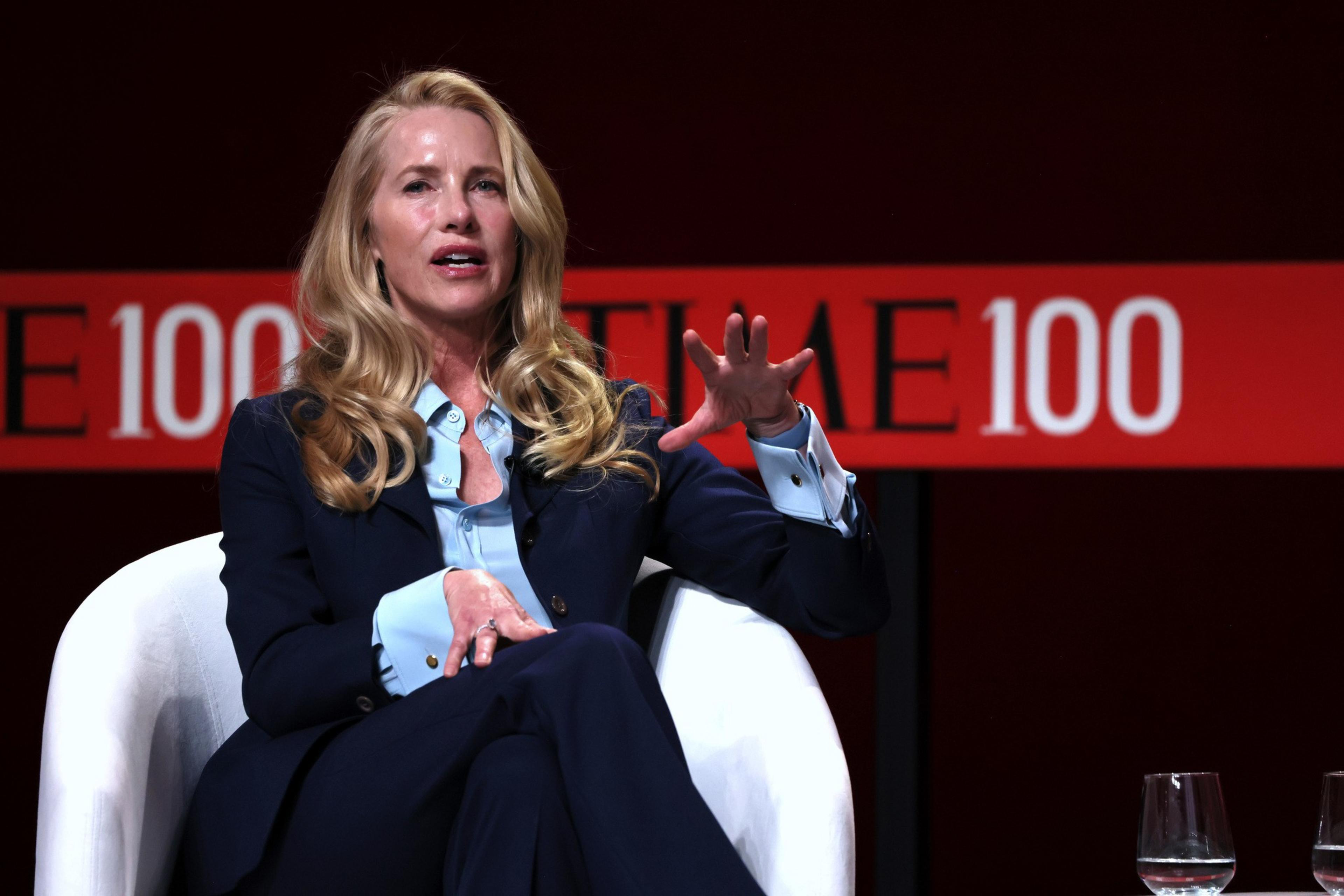 A woman with long blonde hair wearing a navy suit and blue shirt speaks while seated in a white chair, gesturing with her hand. A "TIME 100" banner is in the background.