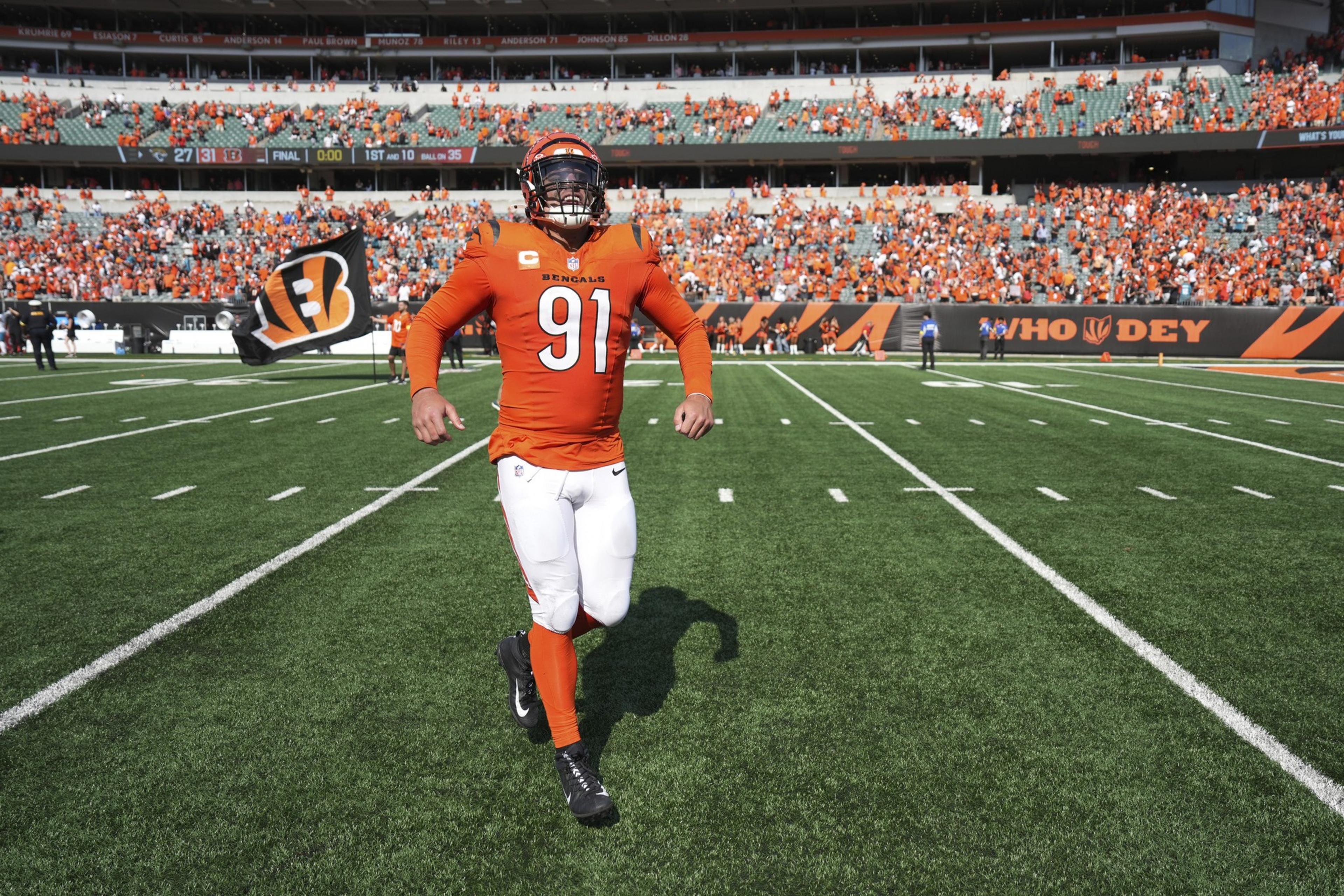 A Cincinnati Bengals football player wearing number 91 runs on the field with a waving Bengals flag behind him and fans in orange filling the stands.