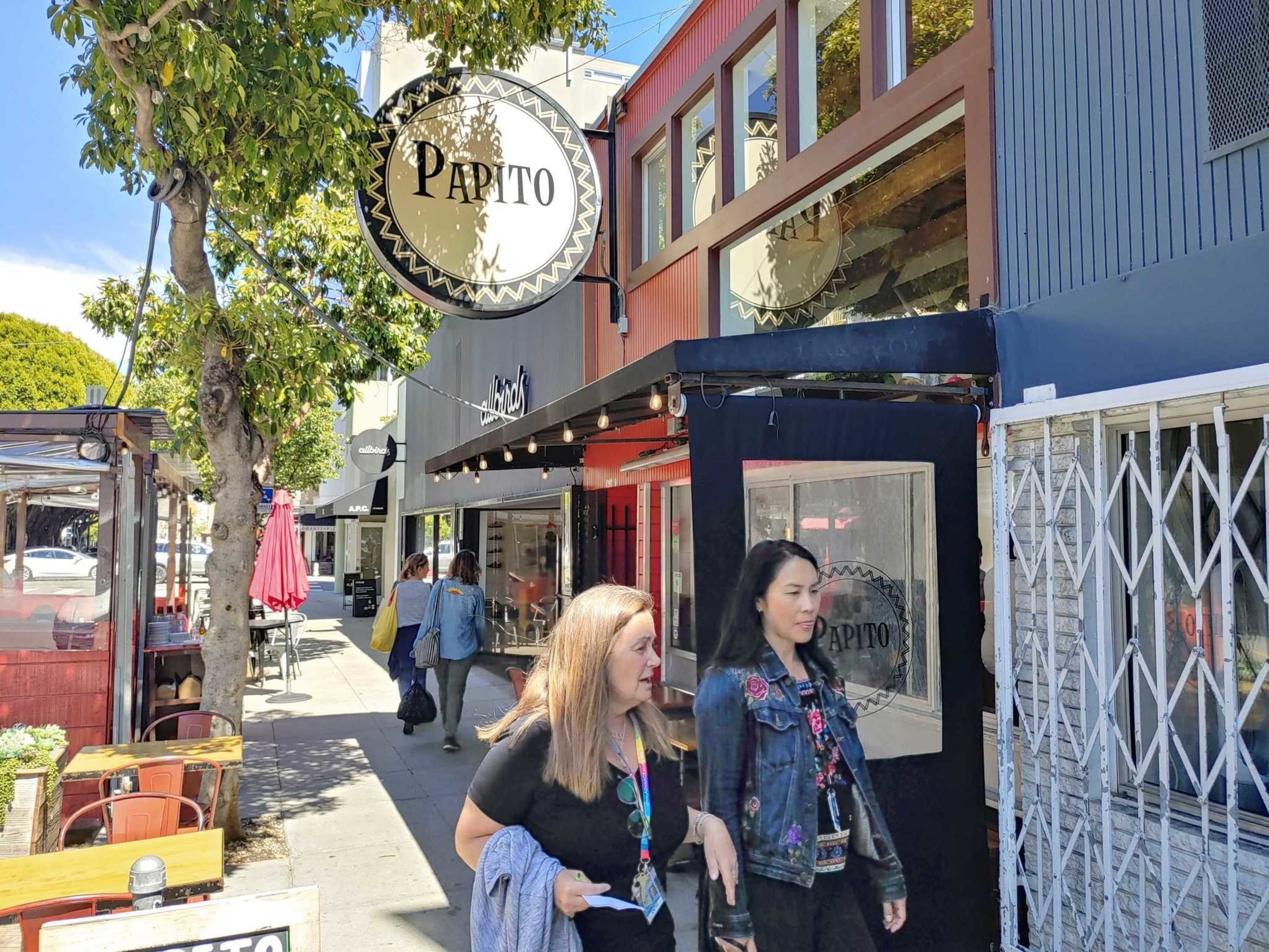 Two women walk past a restaurant named "Papito" on a sunny street. The area has outdoor seating and a tree provides shade, with other people in the background.