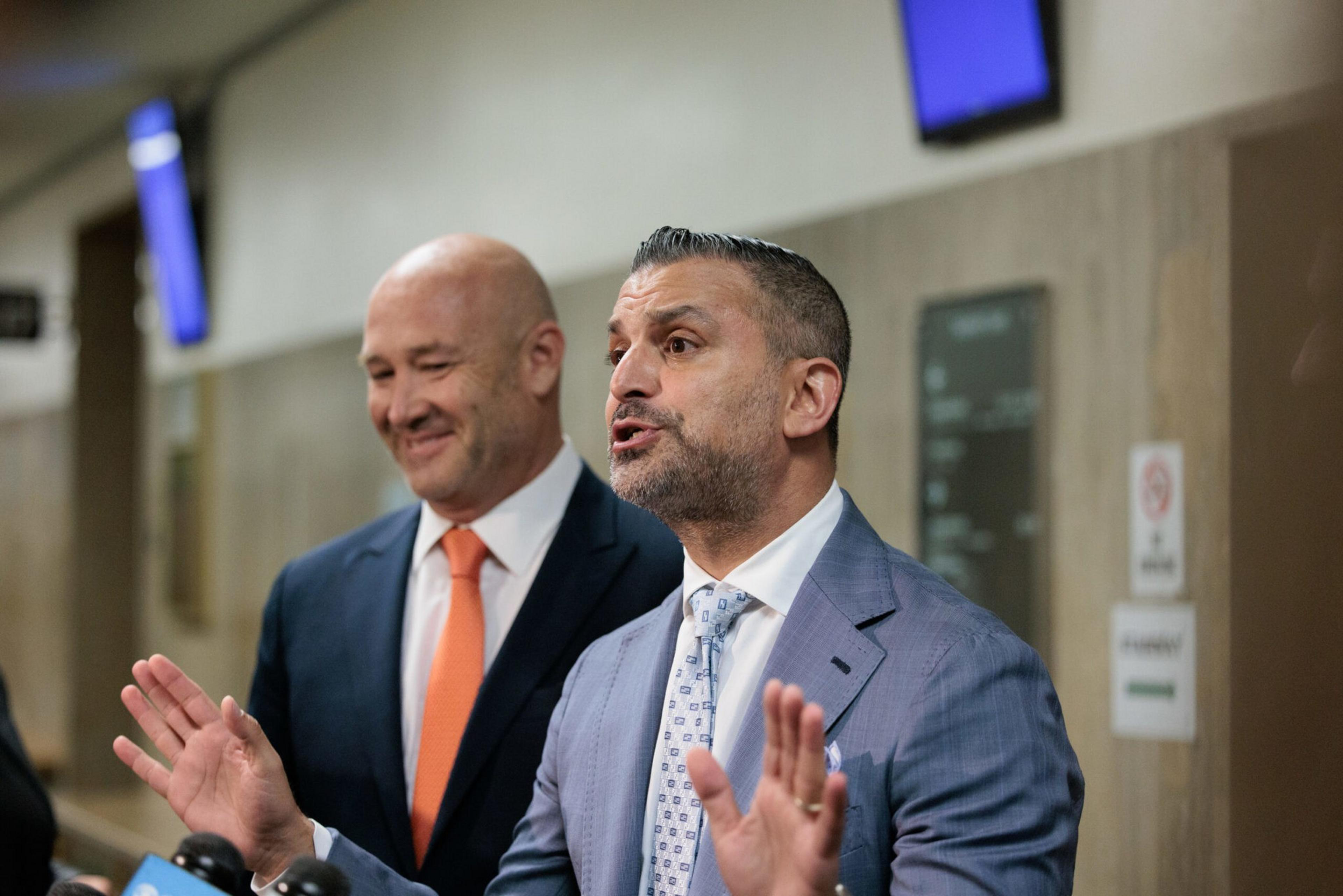 A man in a blue suit and patterned tie gestures while speaking, with another man in a dark suit and orange tie smiling beside him.