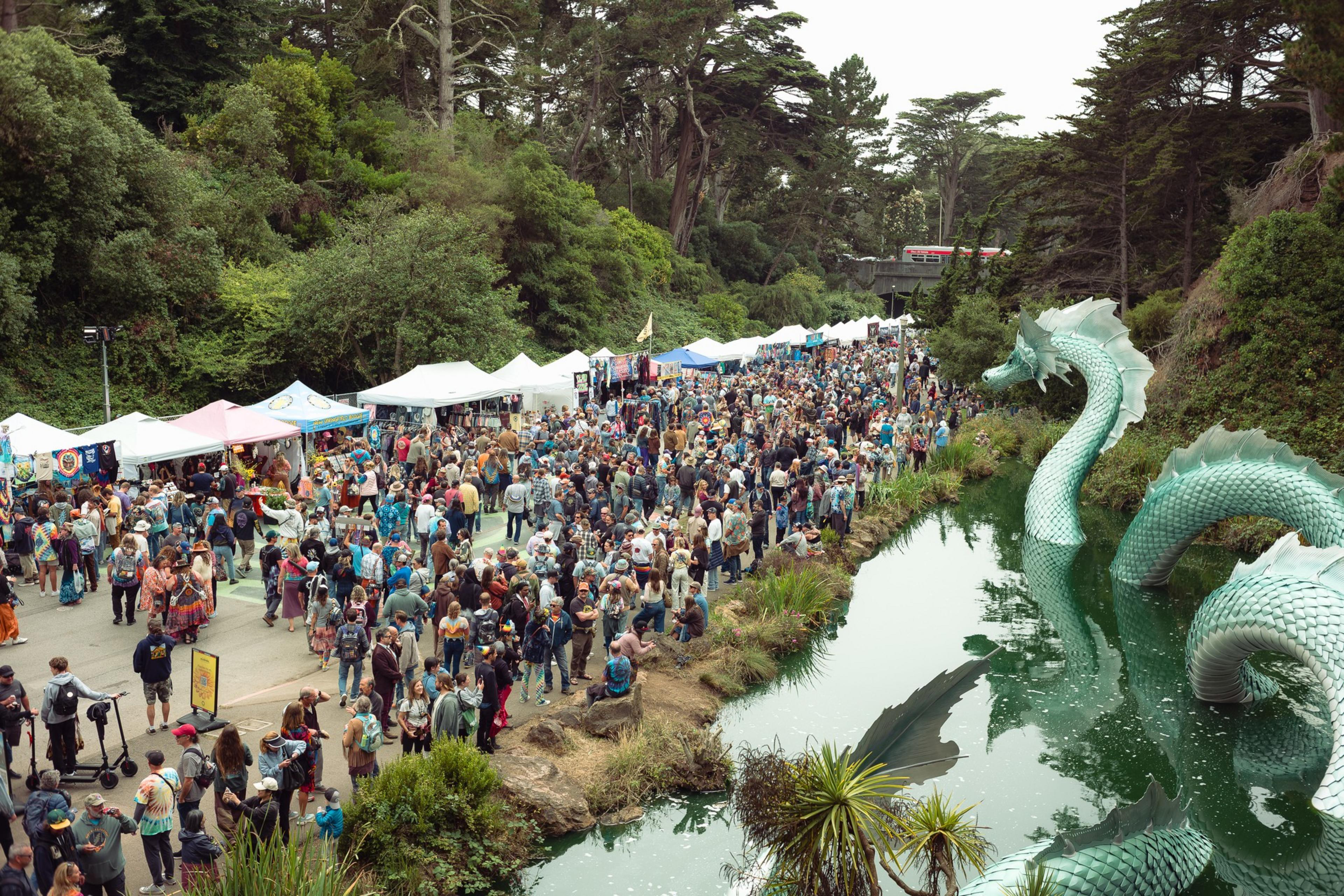 A large crowd gathers along a path lined with white tents near a pond featuring a green, serpentine dragon sculpture.