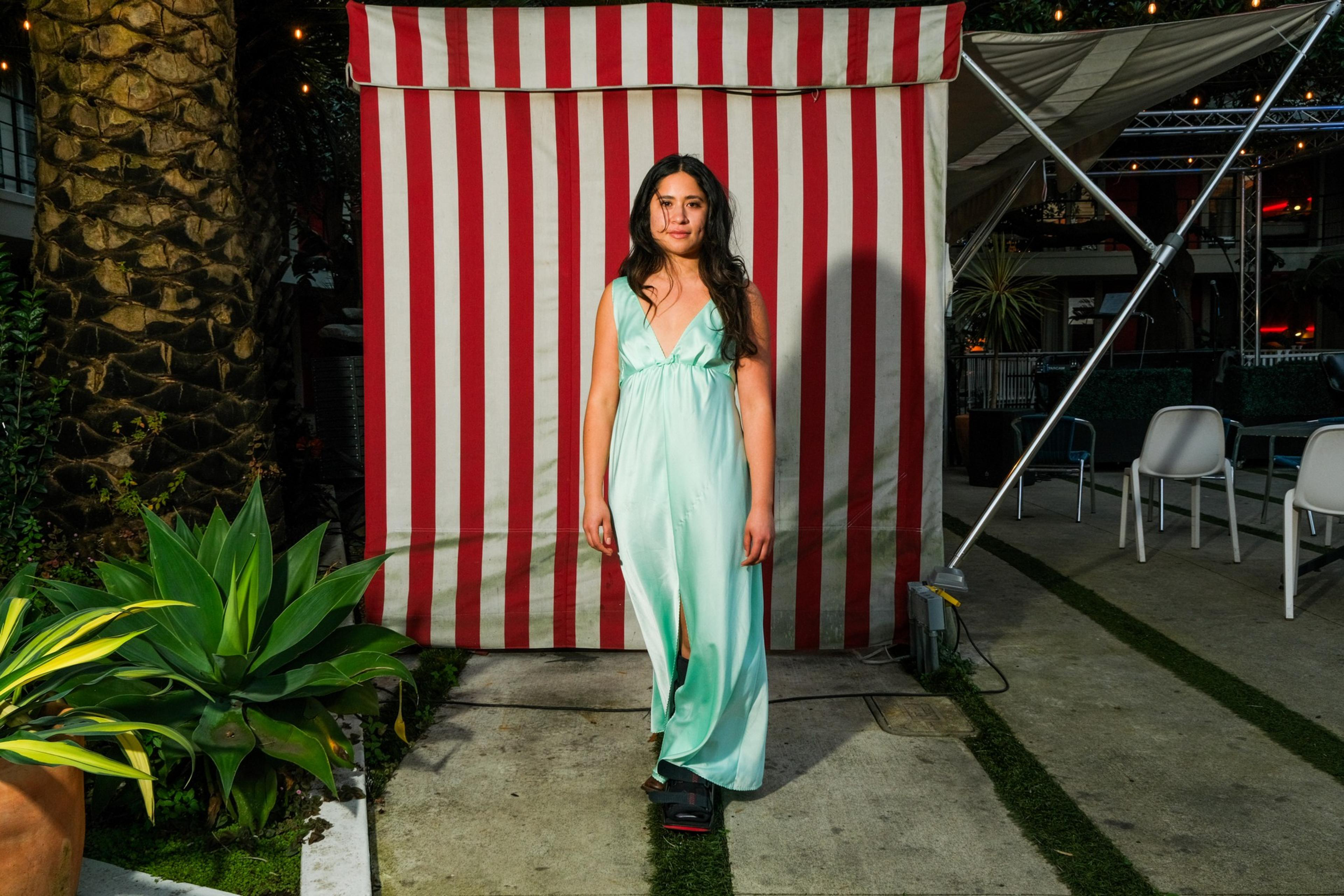 A woman in a light green dress stands in front of a red and white striped backdrop, with plants and outdoor seating nearby.