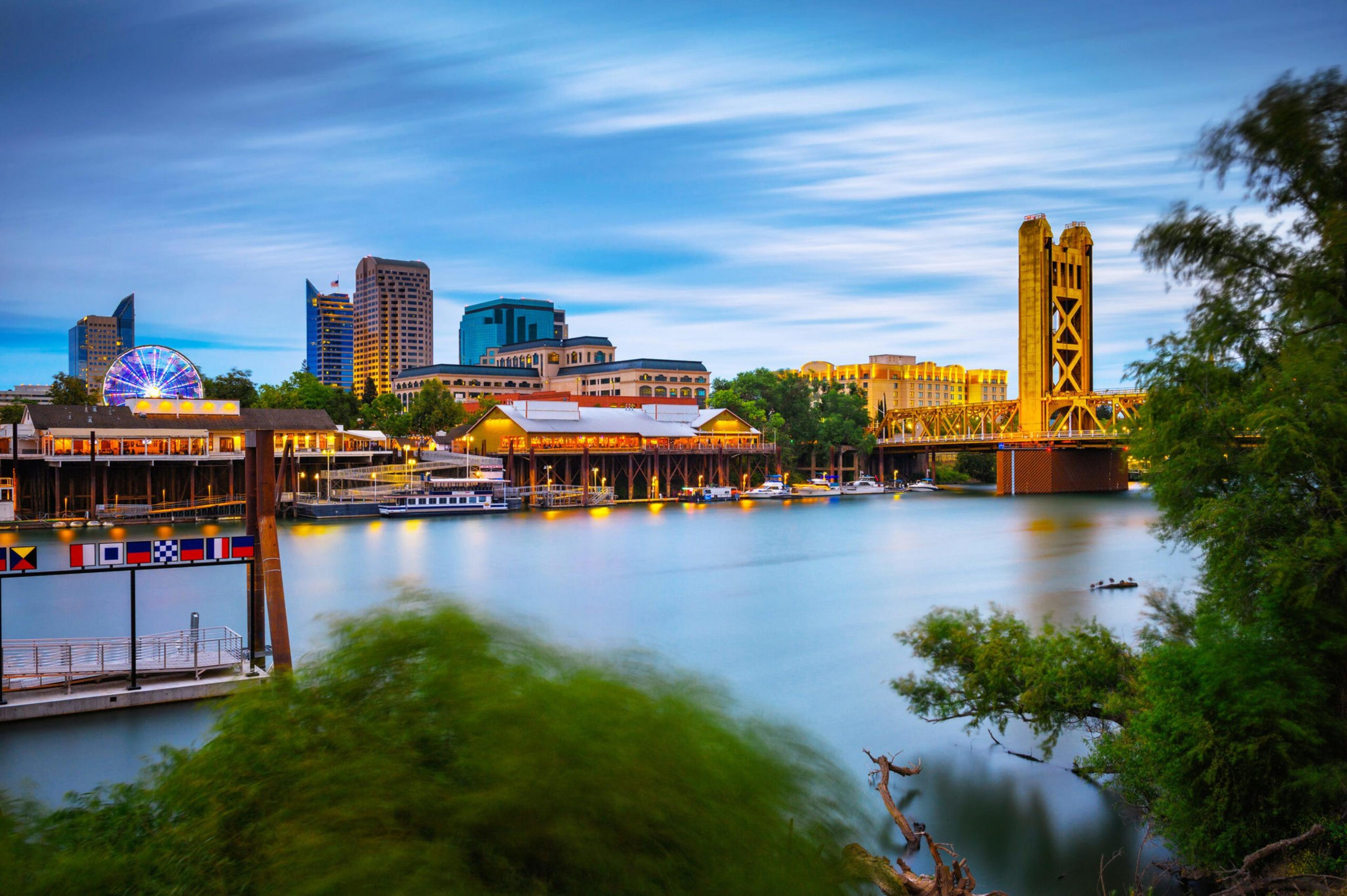 A river reflects city lights with buildings, a Ferris wheel, and a tall illuminated bridge under a blue, partly cloudy sky.