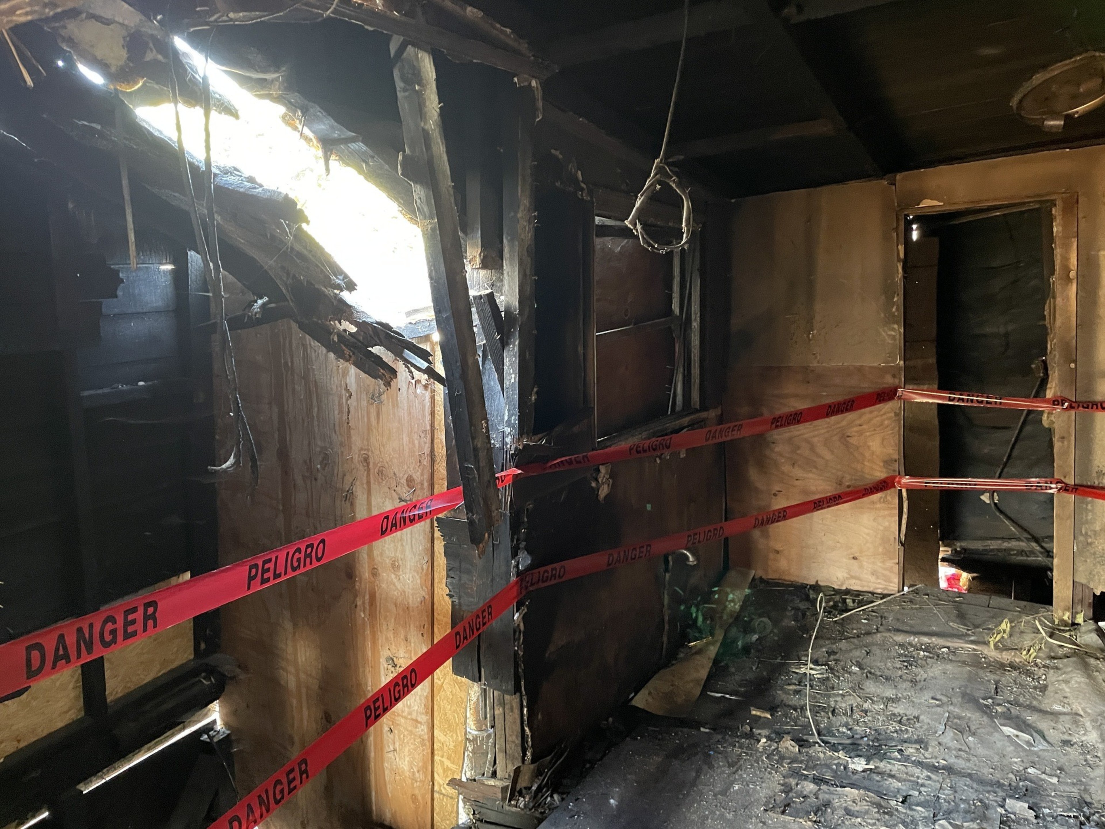 The image shows a charred, damaged room with blackened walls and a partially collapsed ceiling. Red "Danger" tape is strung around, cordoning off the area.
