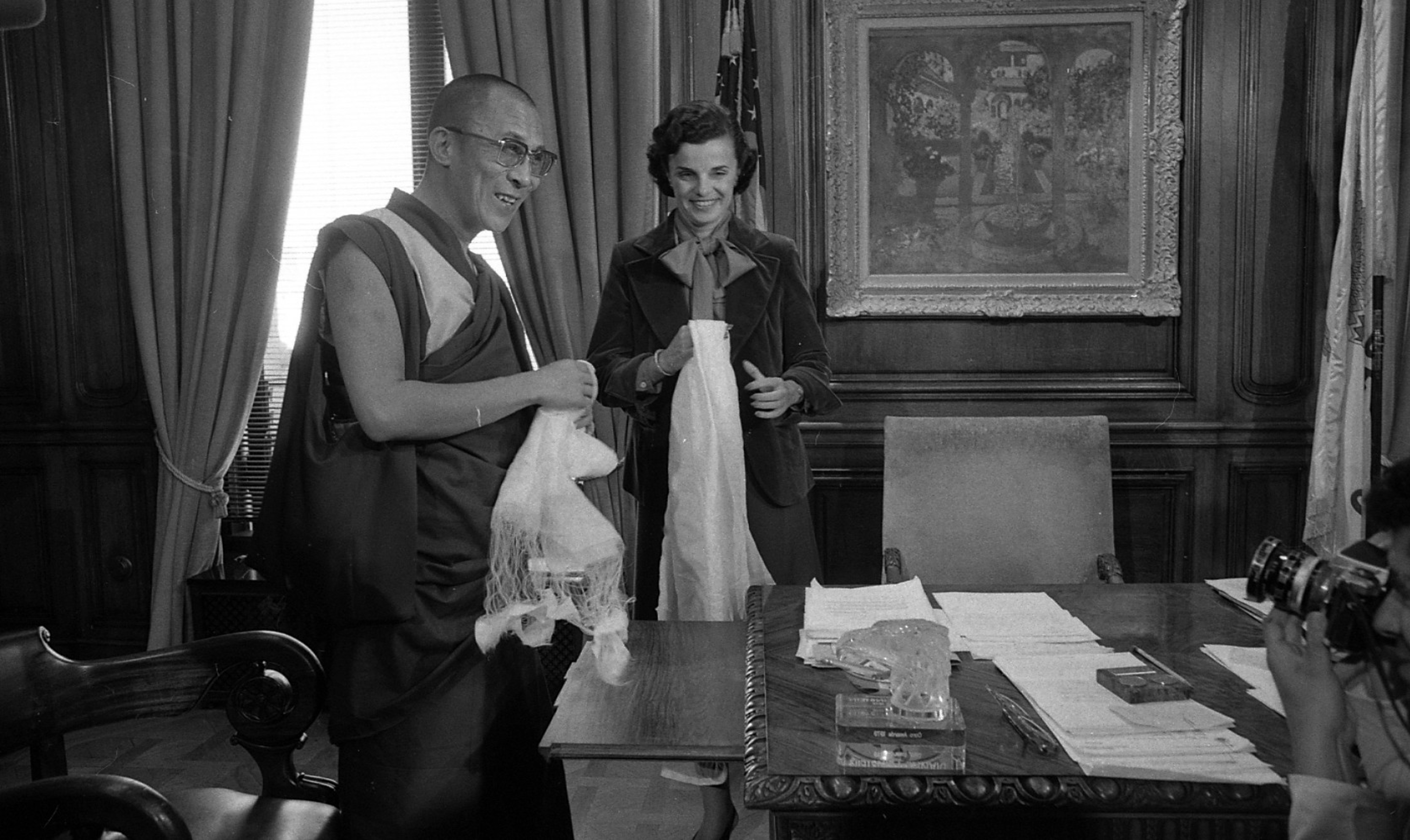 A man in traditional robes and a woman in a suit stand smiling, each holding white scarves, in an ornate room with a desk, papers, and a camera.