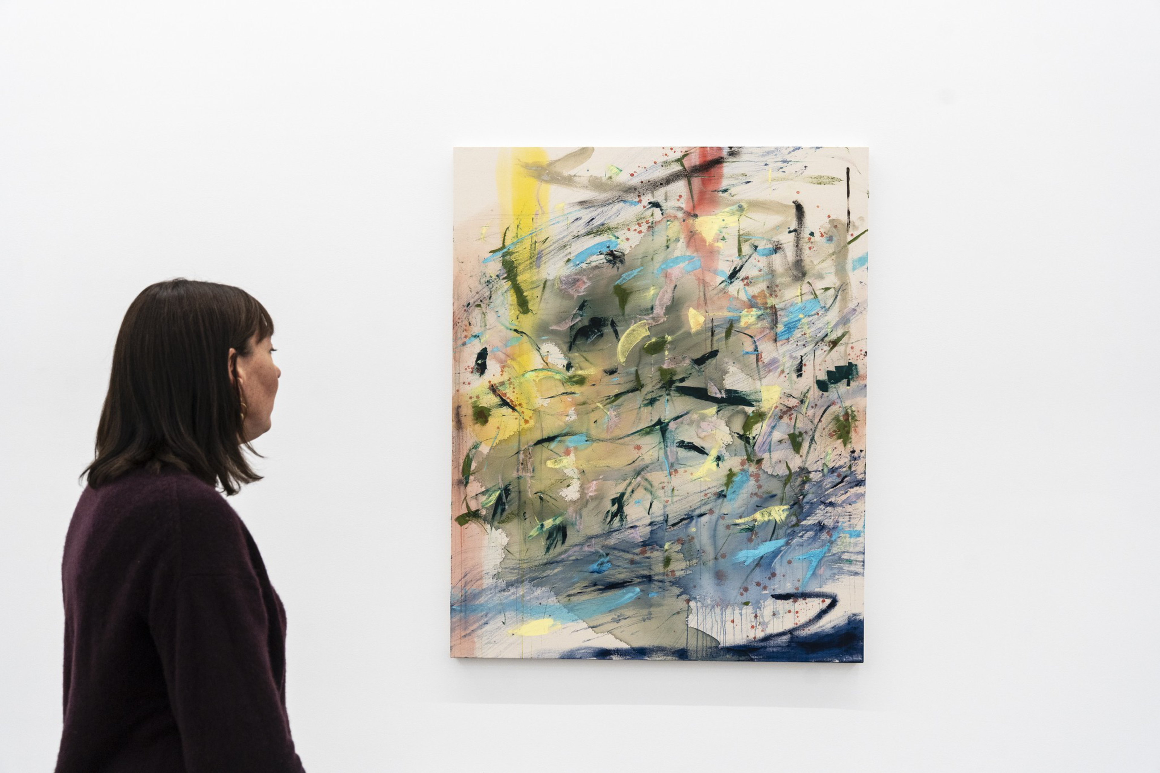 A person with dark hair wearing a dark sweater is looking at a colorful abstract painting with splashes of yellow, blue, green, and black on a white wall.