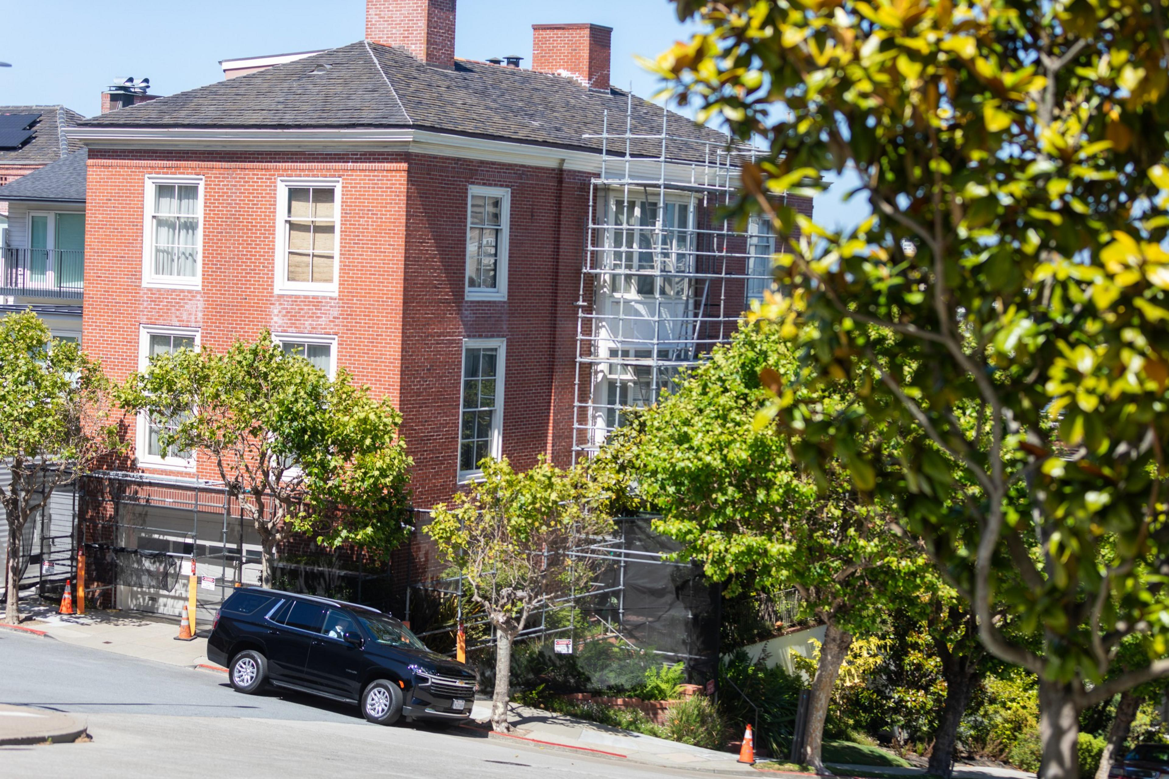 A red-brick building is undergoing maintenance with scaffolding on its side, surrounded by leafy trees and a black SUV parked in front.