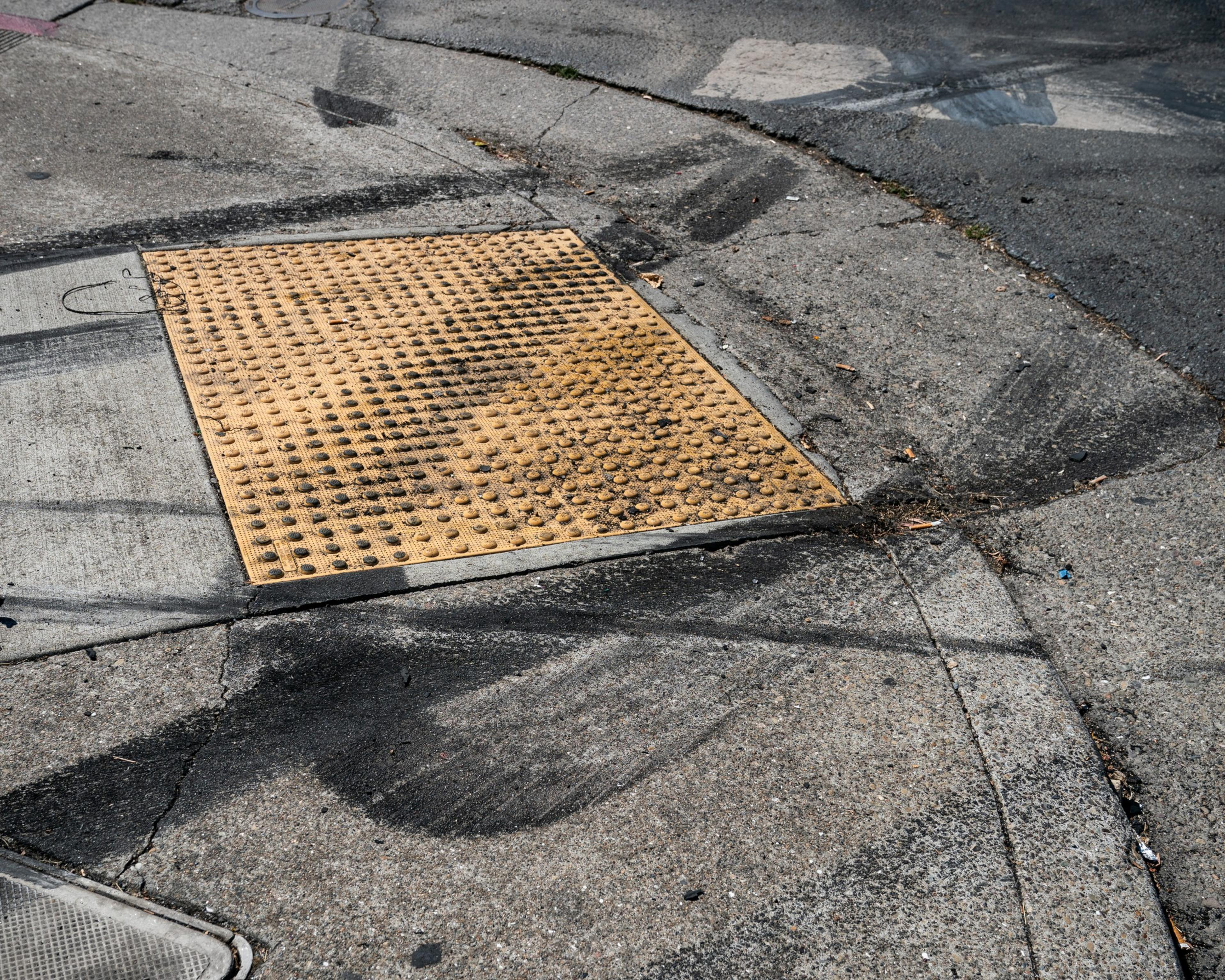 A yellow textured tactile paving patch marks a concrete curb ramp at a street corner, with black tire marks and cracks visible on the surrounding pavement.