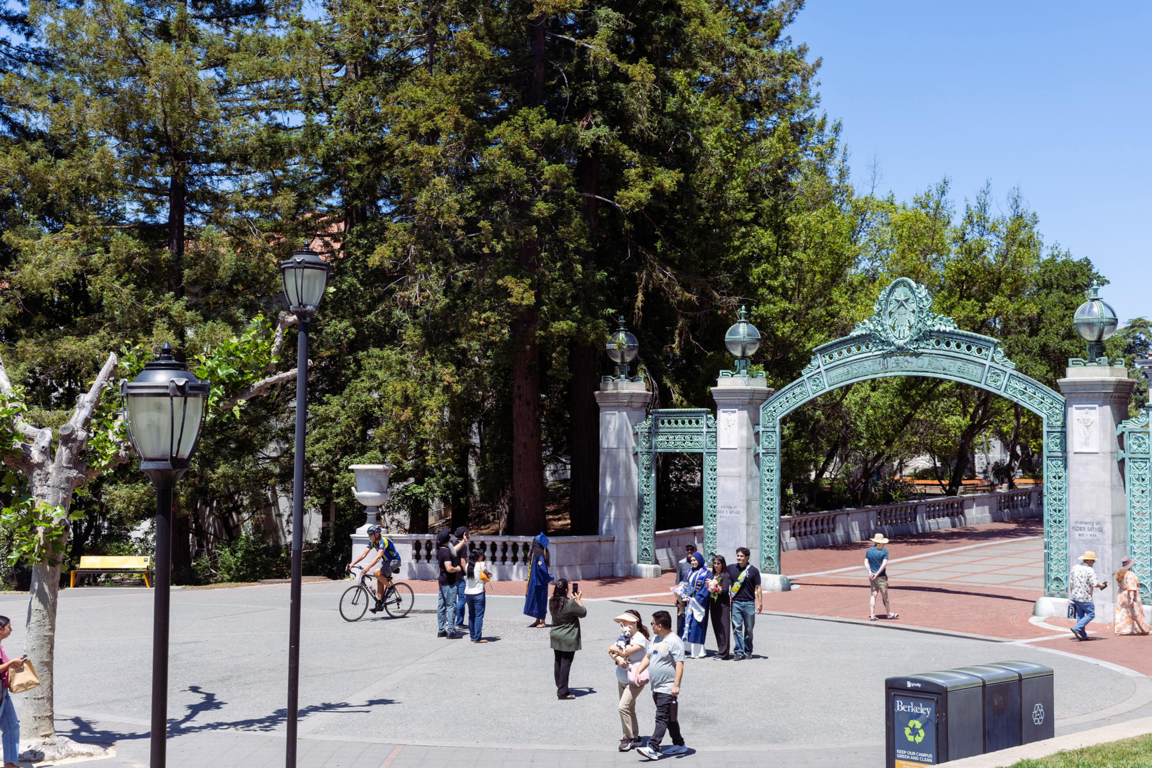 A green ornate archway stands behind a group of people near street lamps, with tall trees and a clear blue sky in the background.