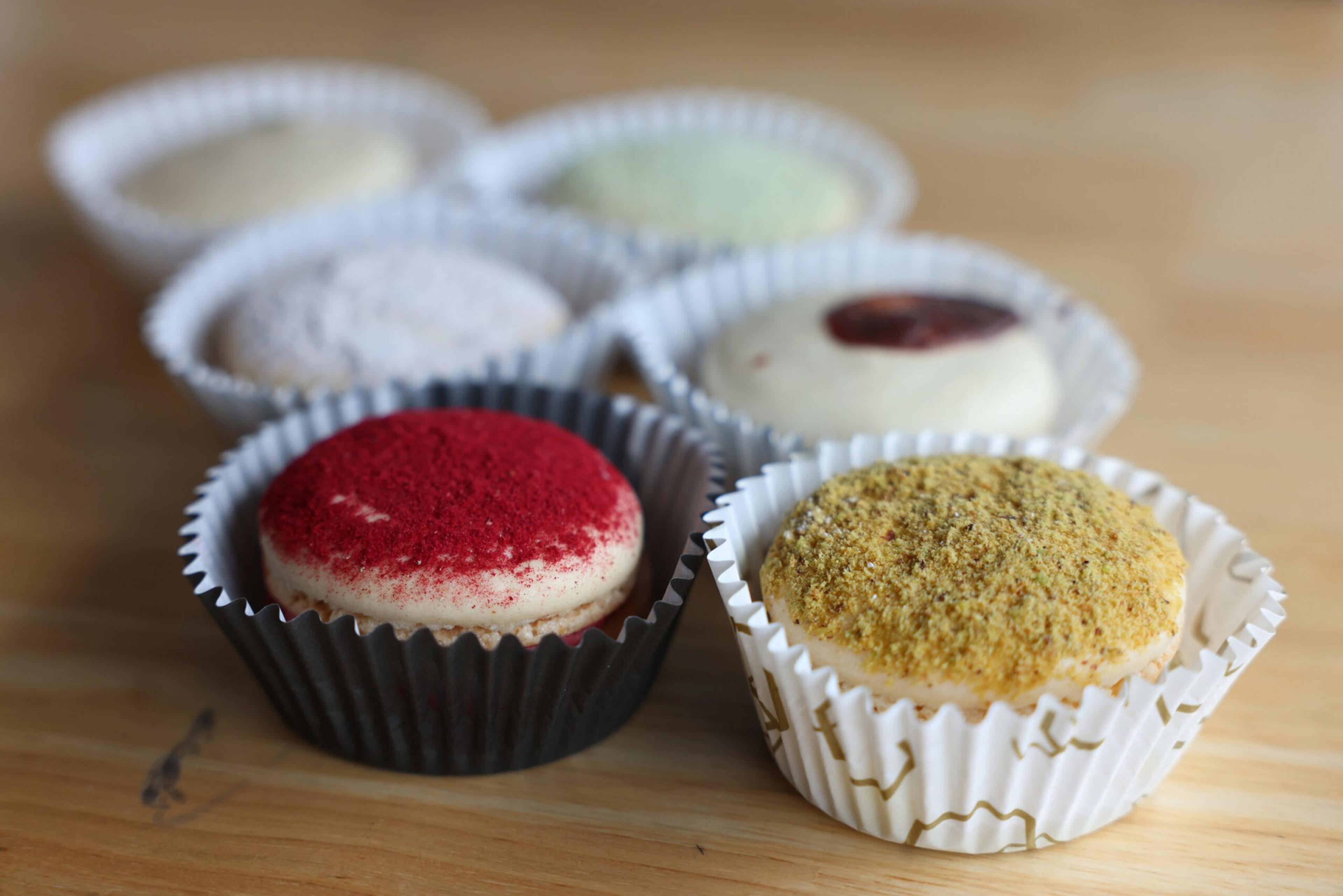 Six assorted macarons sit in individual paper cups on a wooden surface, with toppings like red powder and crushed nuts visible.