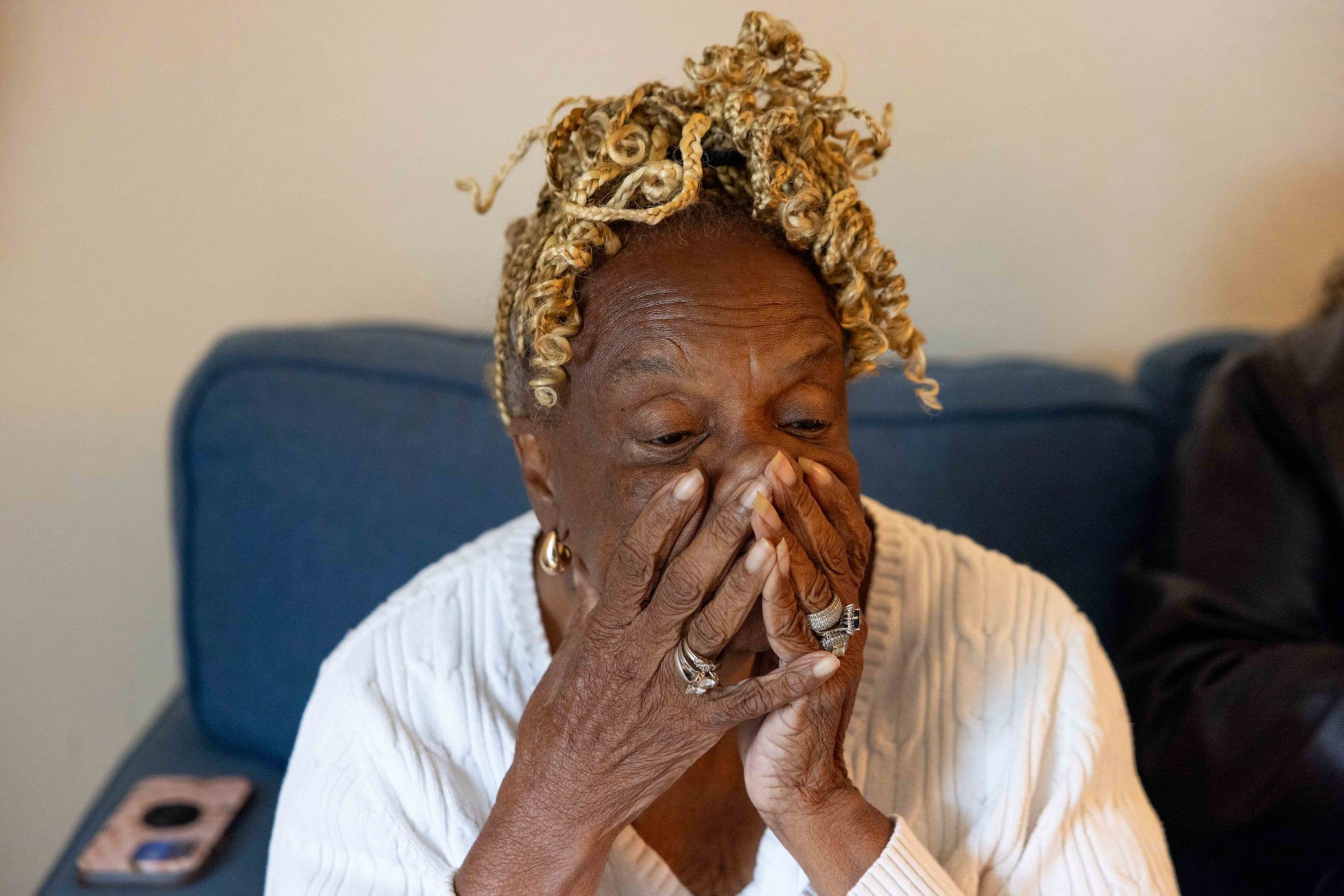 An elderly woman with curly blonde hair covers her mouth with both hands, sitting on a blue couch, wearing a white sweater and multiple rings.