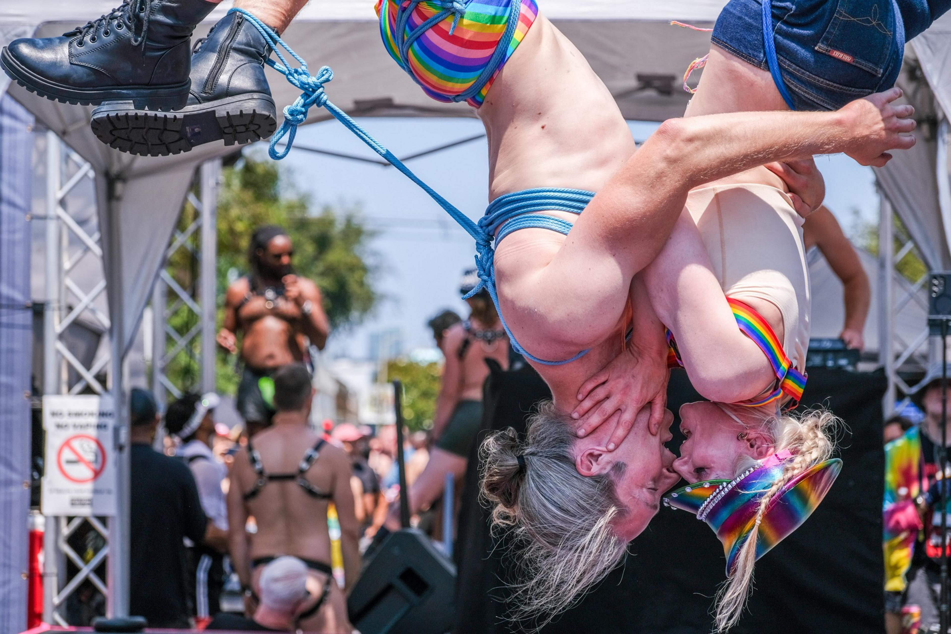Two people are suspended upside down, embracing each other. They are wearing colorful rainbow attire and surrounded by a crowd, possibly at a festival or event.