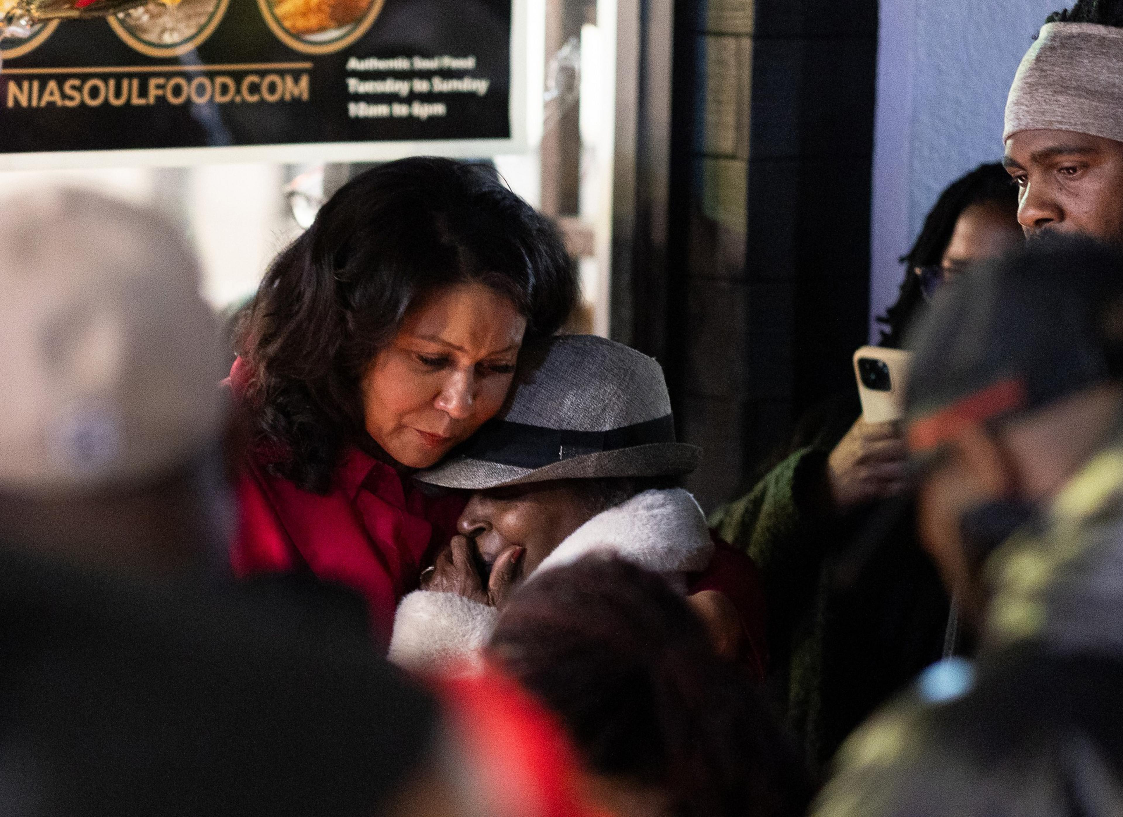 A woman in a red coat is comforting another, who is wearing a gray hat, amidst a group. They're standing outside a building, with an emotional atmosphere.