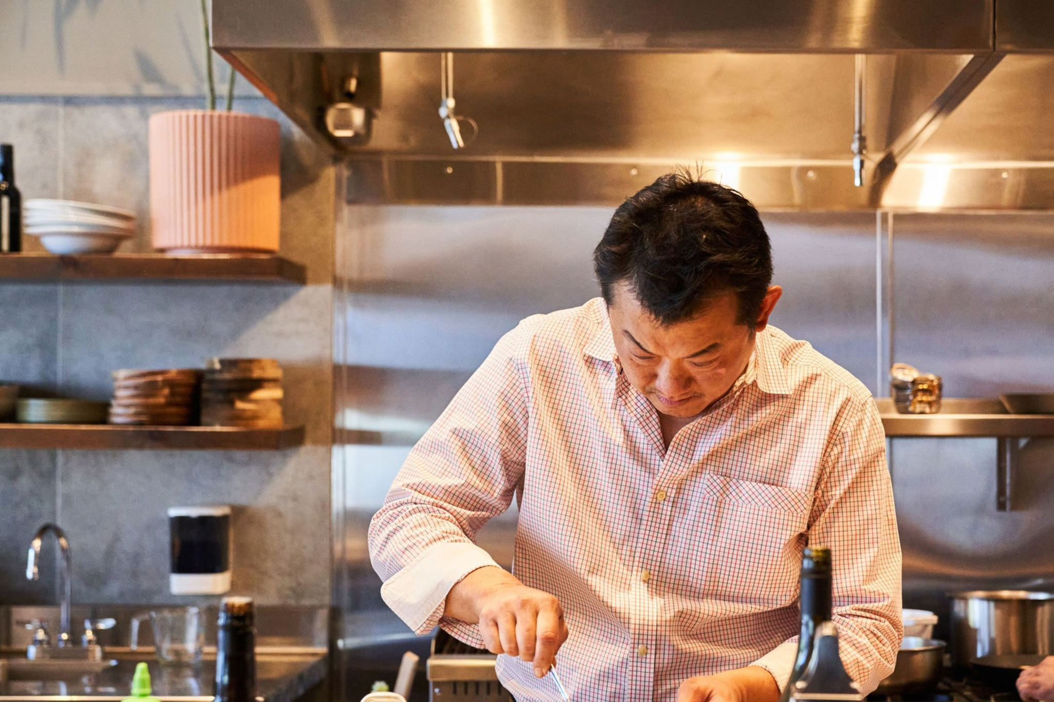 Two chefs are in a professional kitchen. One is focusing on mixing ingredients in a bowl, while the other is cooking at the stove. Shelves with dishes are visible.
