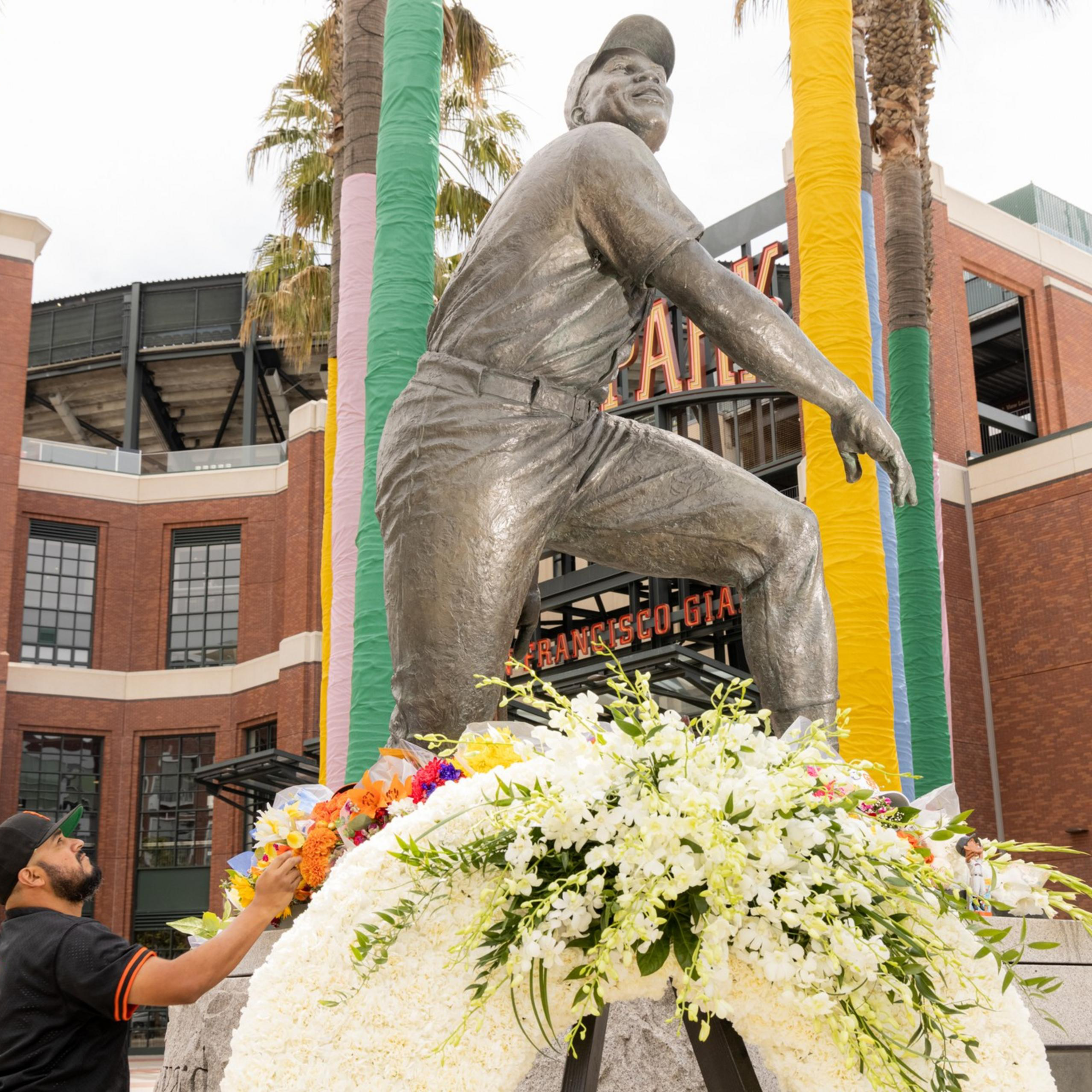 A statue of a baseball player is surrounded by colorful wrapped poles and flowers. A man wearing a baseball jersey is adjusting a floral display at the statue's base.