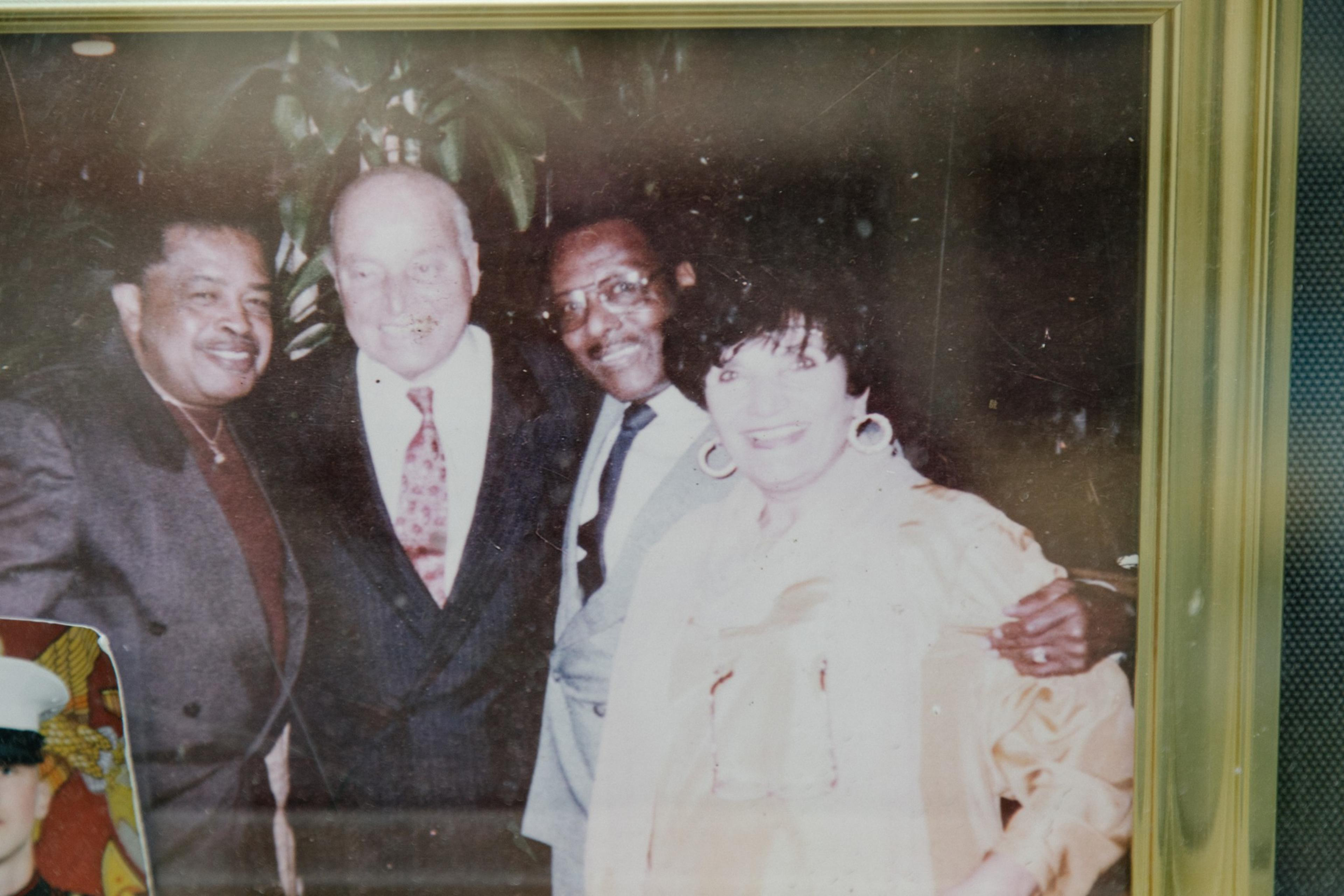 Four people posing for a photo, two men on the left, a woman on the right, with another man partly behind her. They are smiling, dressed in formal wear.