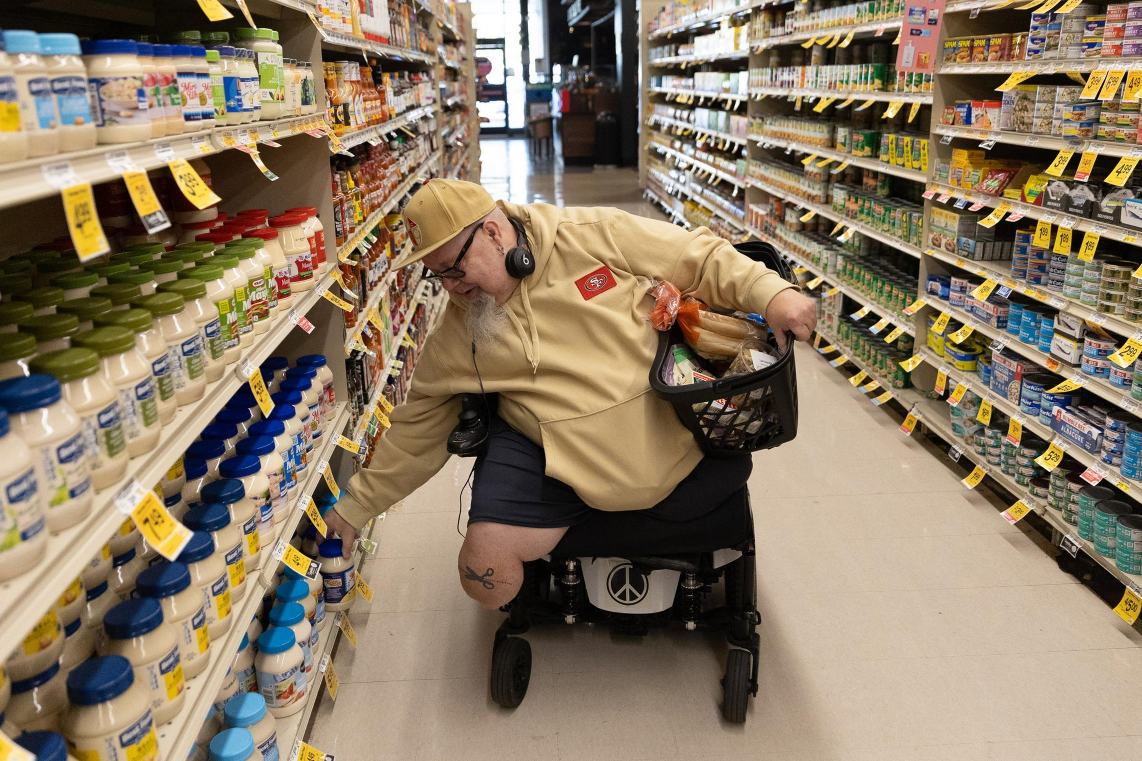 A person in a motorized wheelchair wearing headphones is shopping in a grocery aisle. They reach for a jar, holding a basket filled with items.
