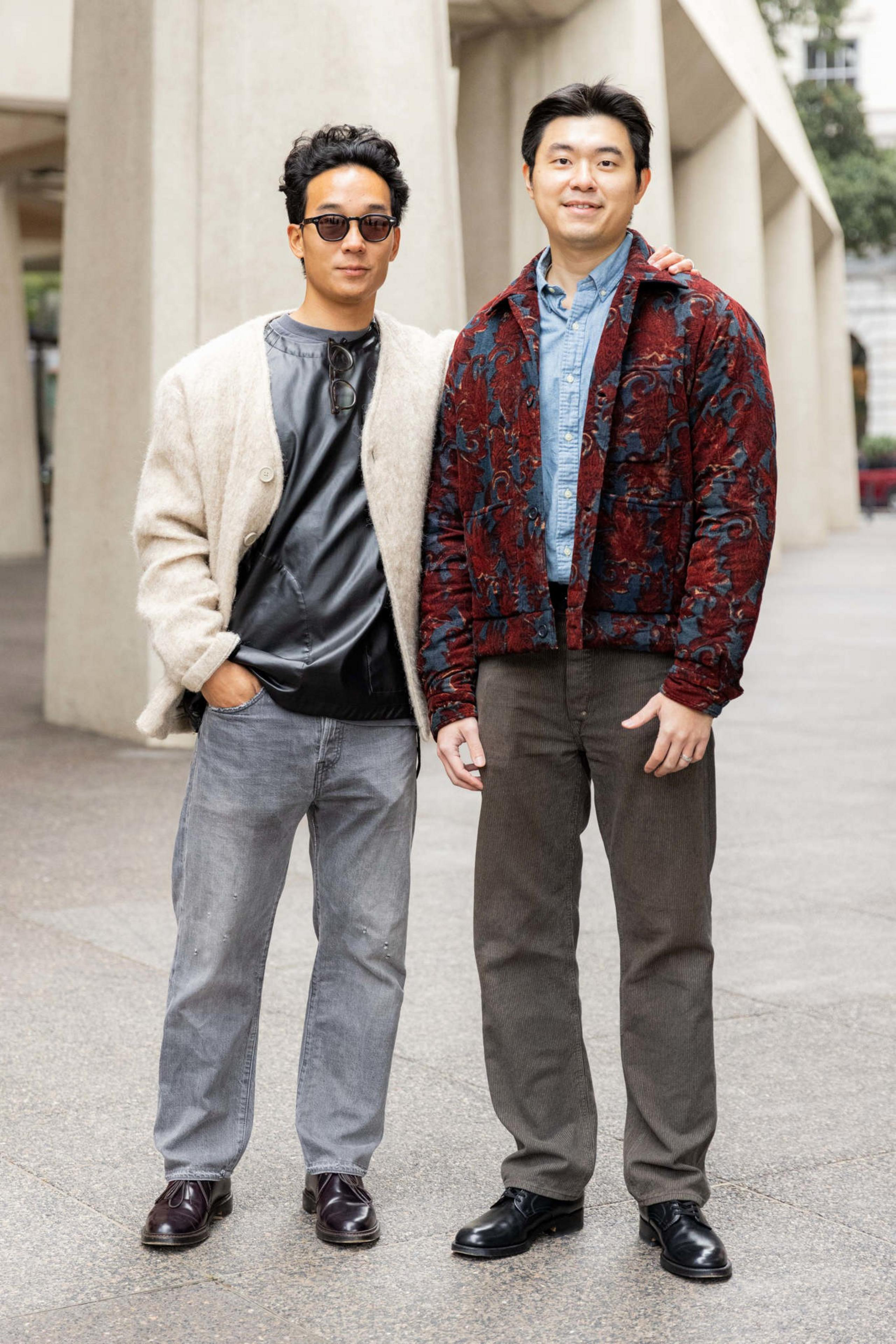 Two men stand side by side outdoors; one wears sunglasses, a beige jacket, and gray jeans, while the other wears a red patterned jacket over a blue shirt and dark pants.