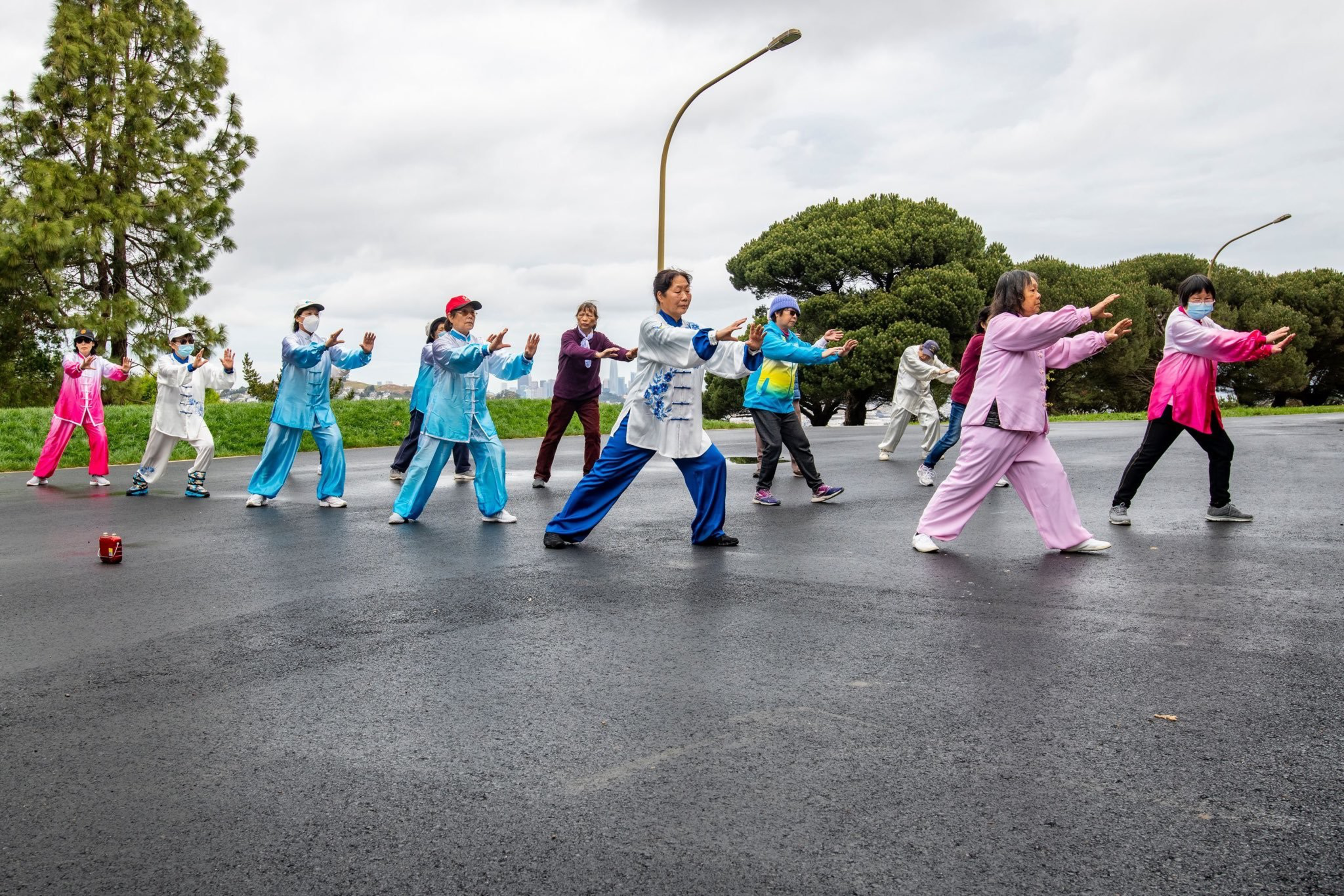 A group of people dressed in colorful clothes practice Tai Chi outdoors on a paved area with trees and cloudy sky in the background.