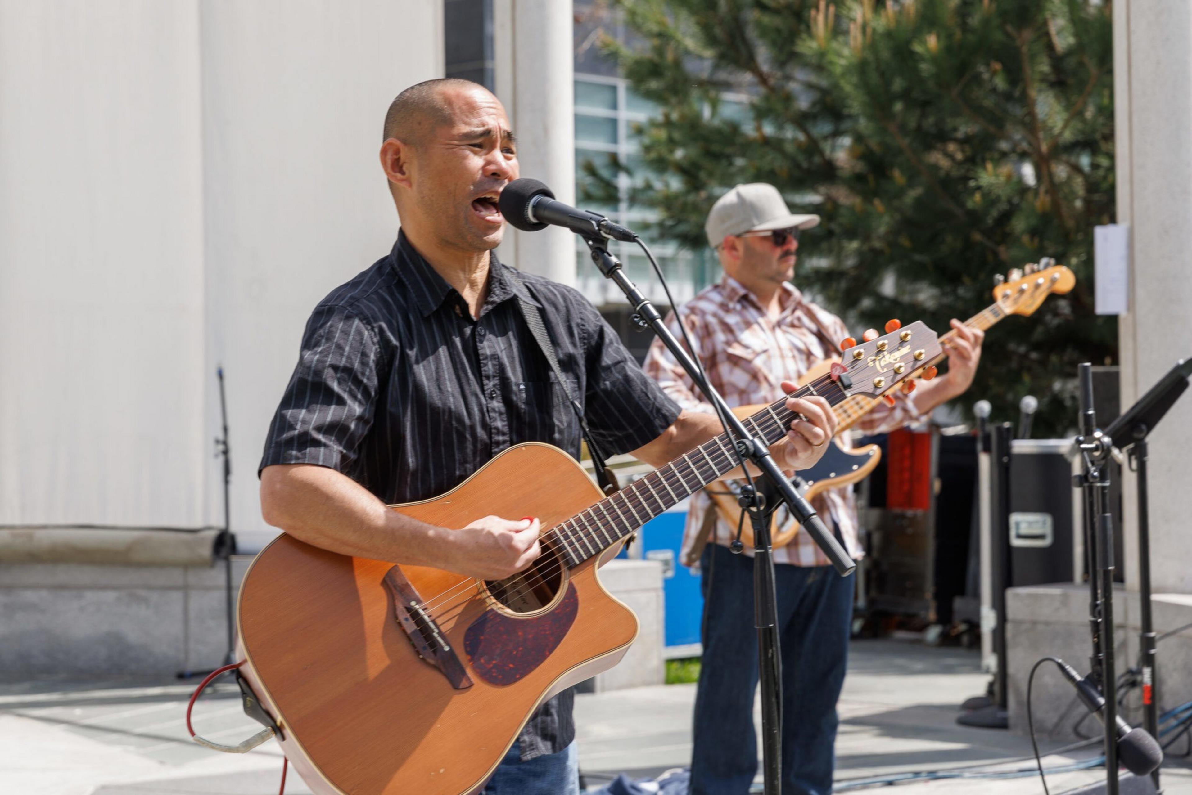 A man is singing and playing an acoustic guitar, with another playing an electric guitar, both performing outdoors.