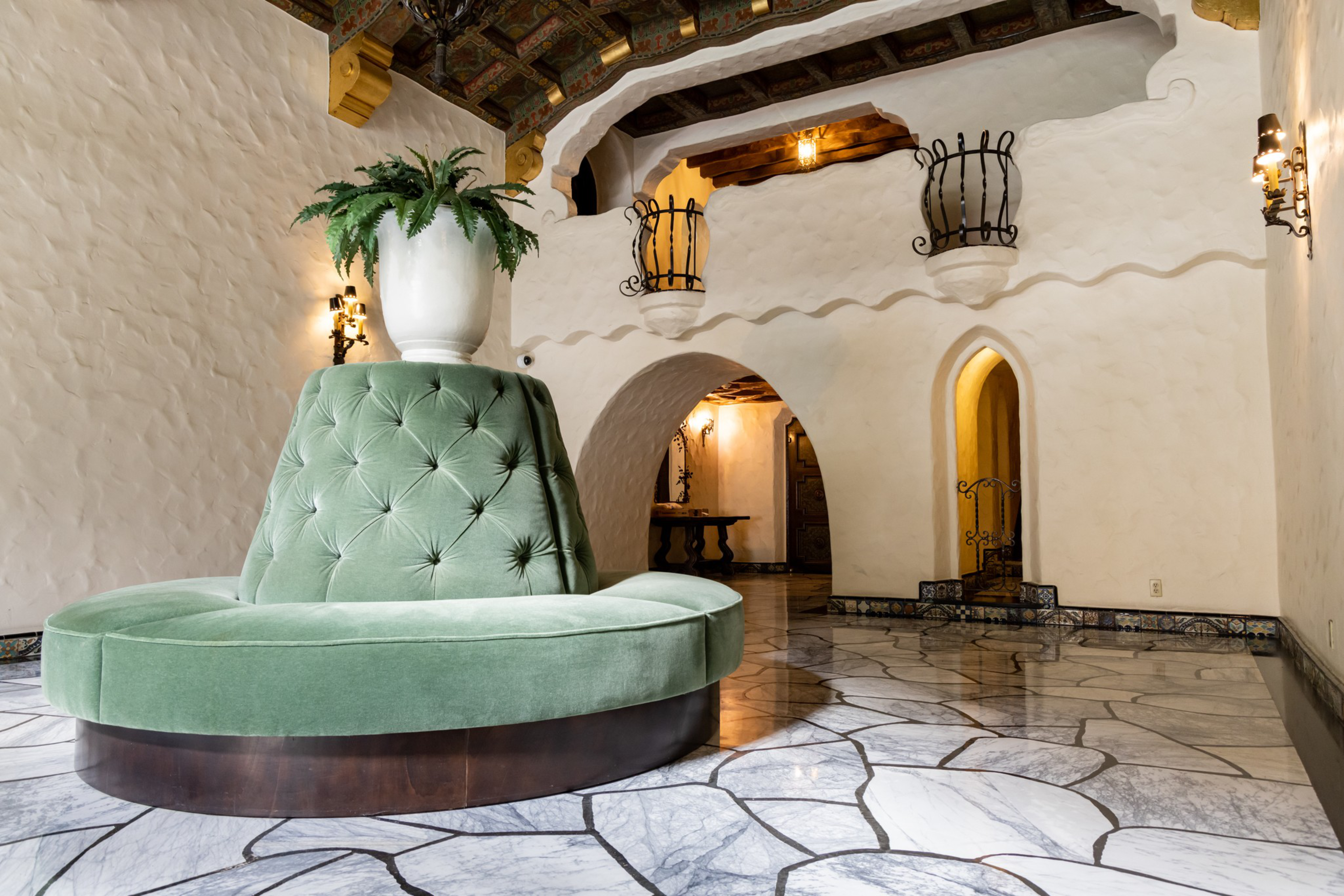 A spacious room with a tufted green circular bench surrounding a vase of greenery, white textured walls, arched doorways, and a patterned marble floor.