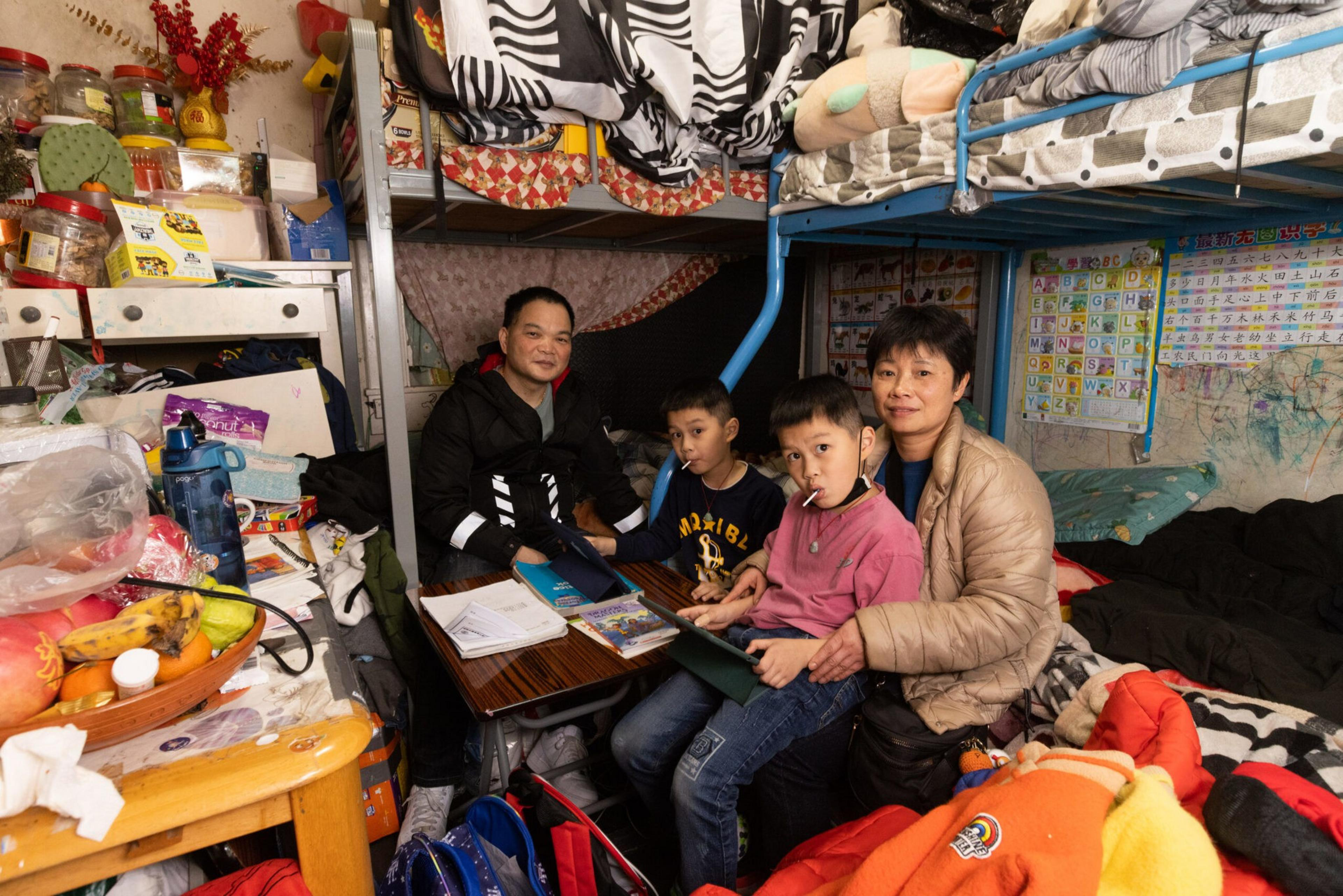 A family of four sits closely in a cluttered room with bunk beds, study materials, and colorful posters on the wall behind them.
