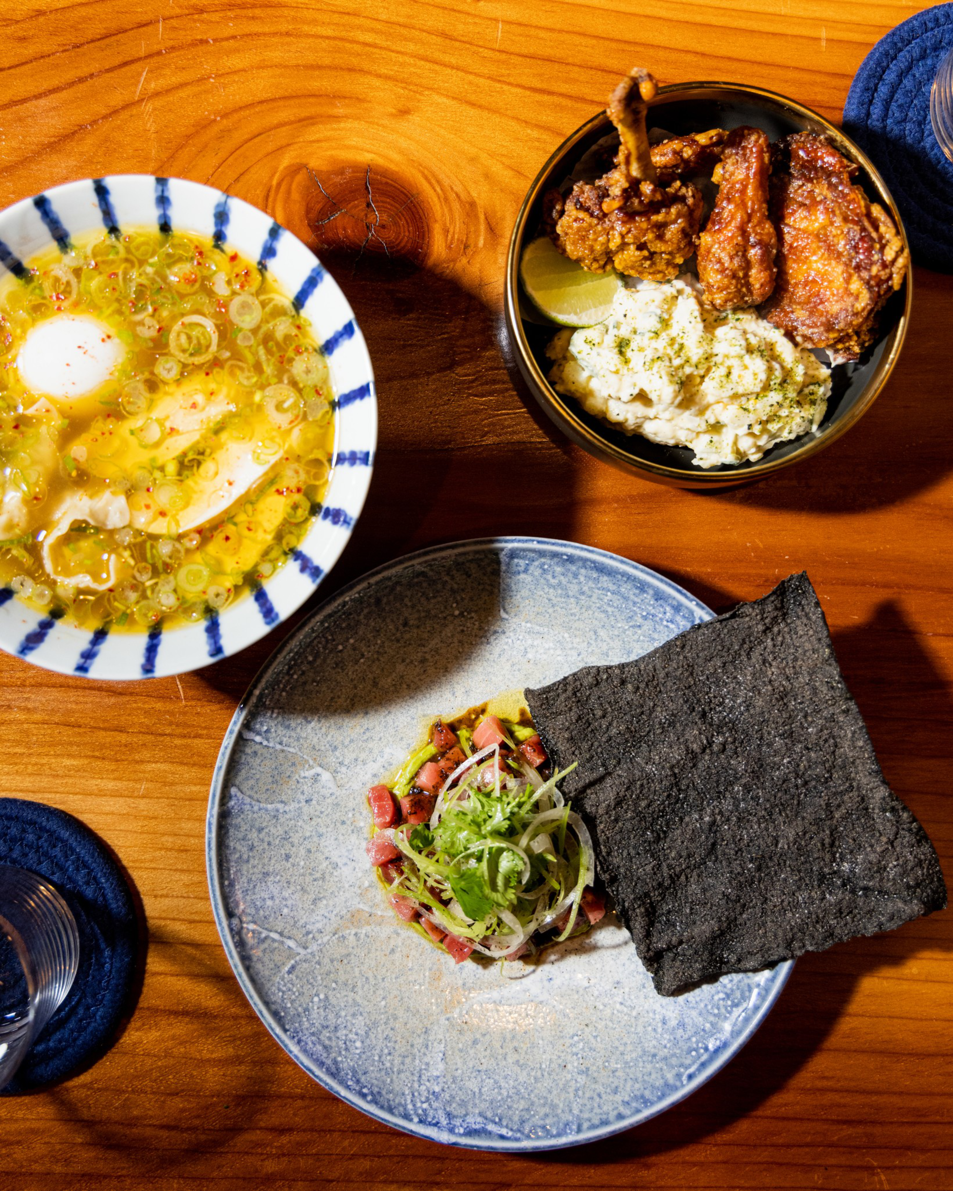 A bowl of soup with egg and scallions, a bowl with fried chicken and potato salad, and a plate with diced meat, greens, and two large black crackers.
