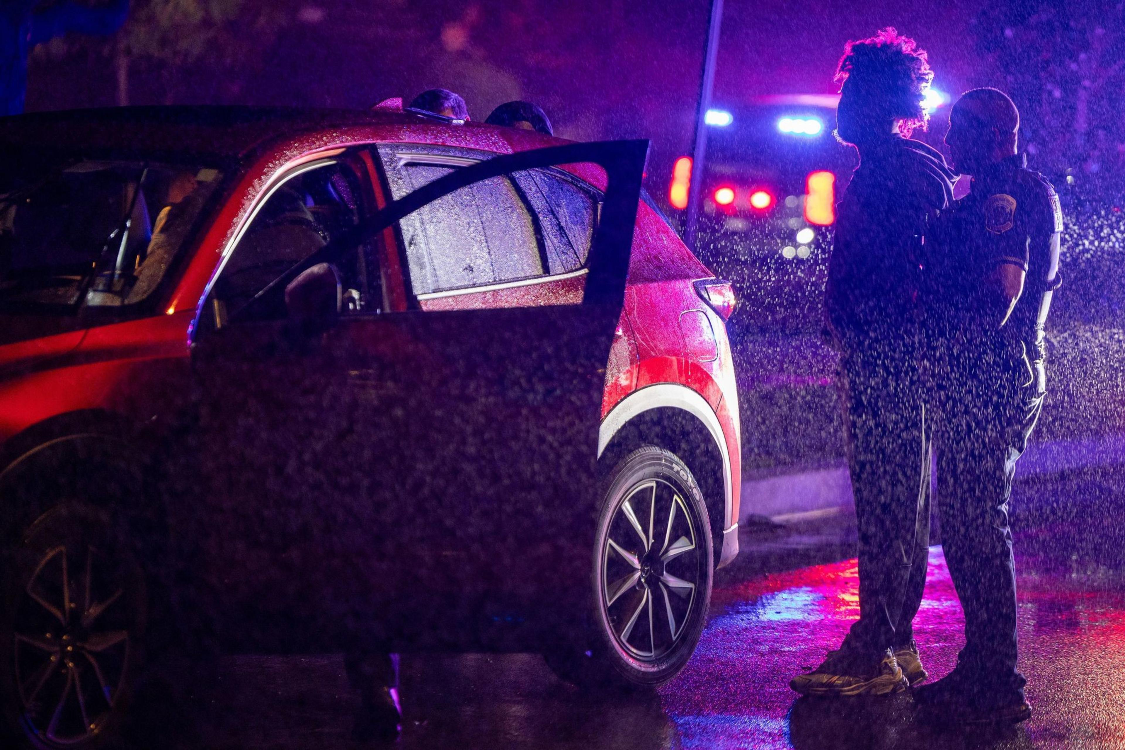 A person is being handcuffed by a police officer next to a red SUV with its door open during a rainy night with flashing police lights.