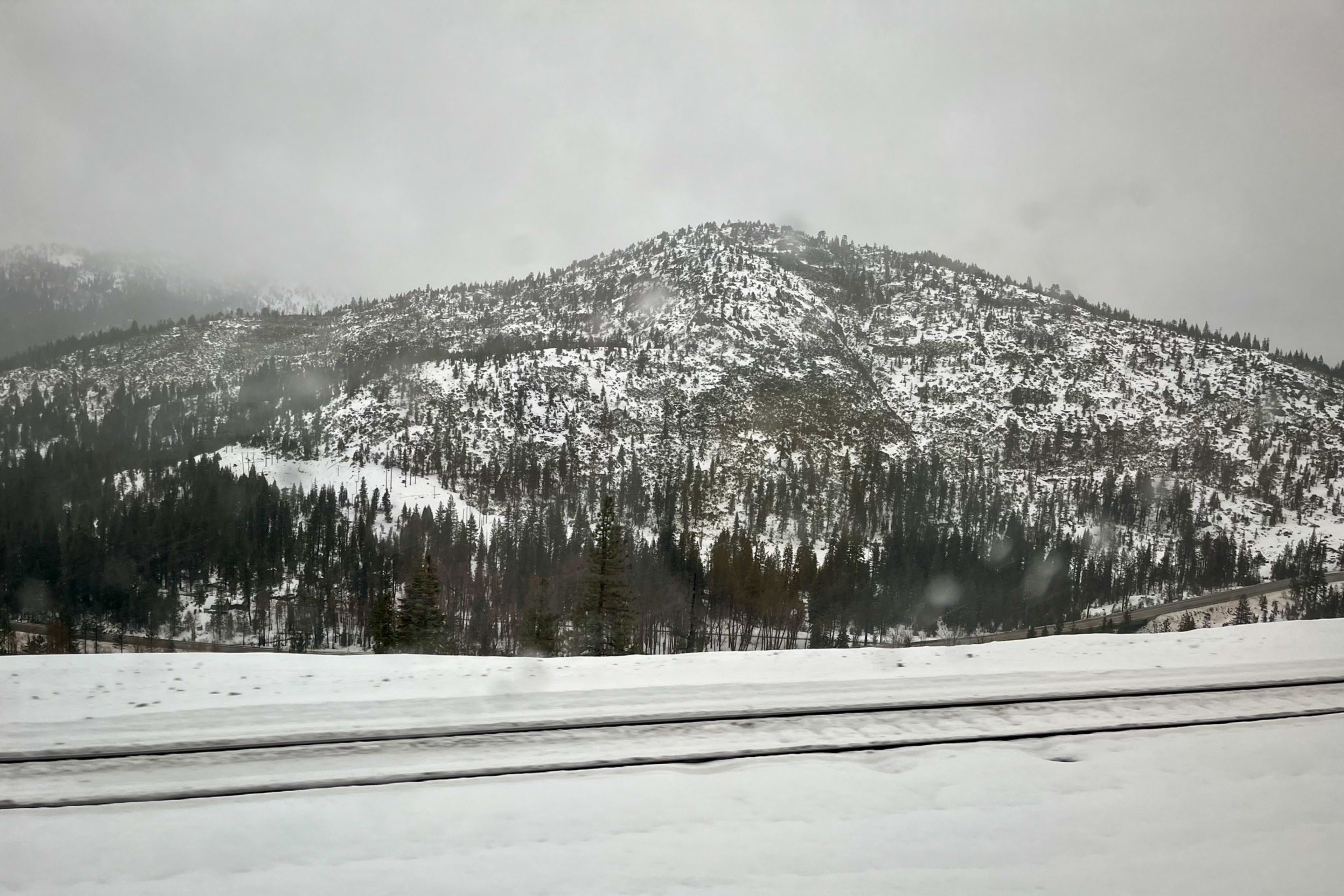 Snow-covered railroad tracks run parallel in front of a forested mountain dusted with snow under a gray, overcast sky.