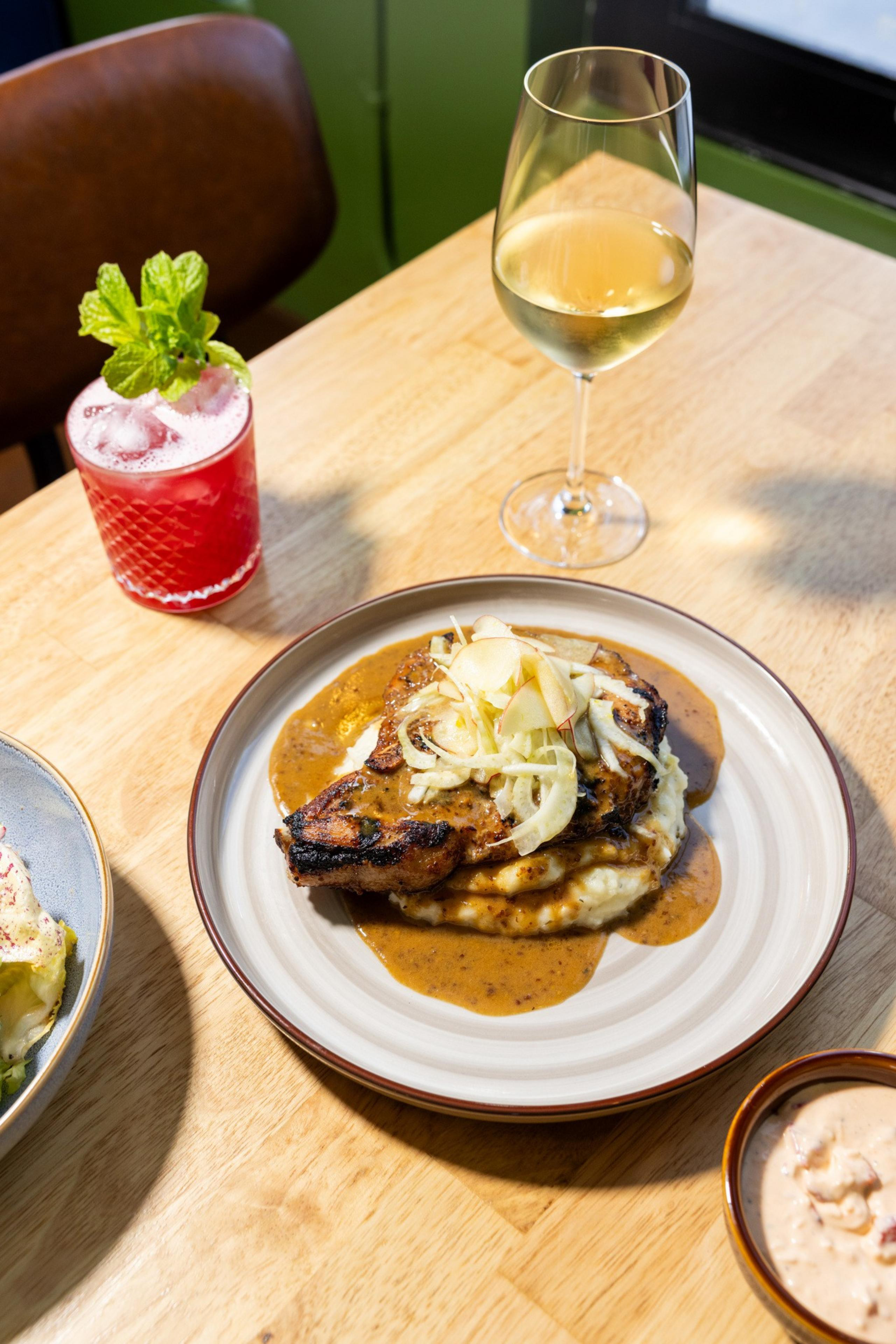 A delicious meal featuring grilled meat on creamy mashed potatoes with sauce, topped with sliced vegetables, accompanied by a cocktail and a glass of white wine.