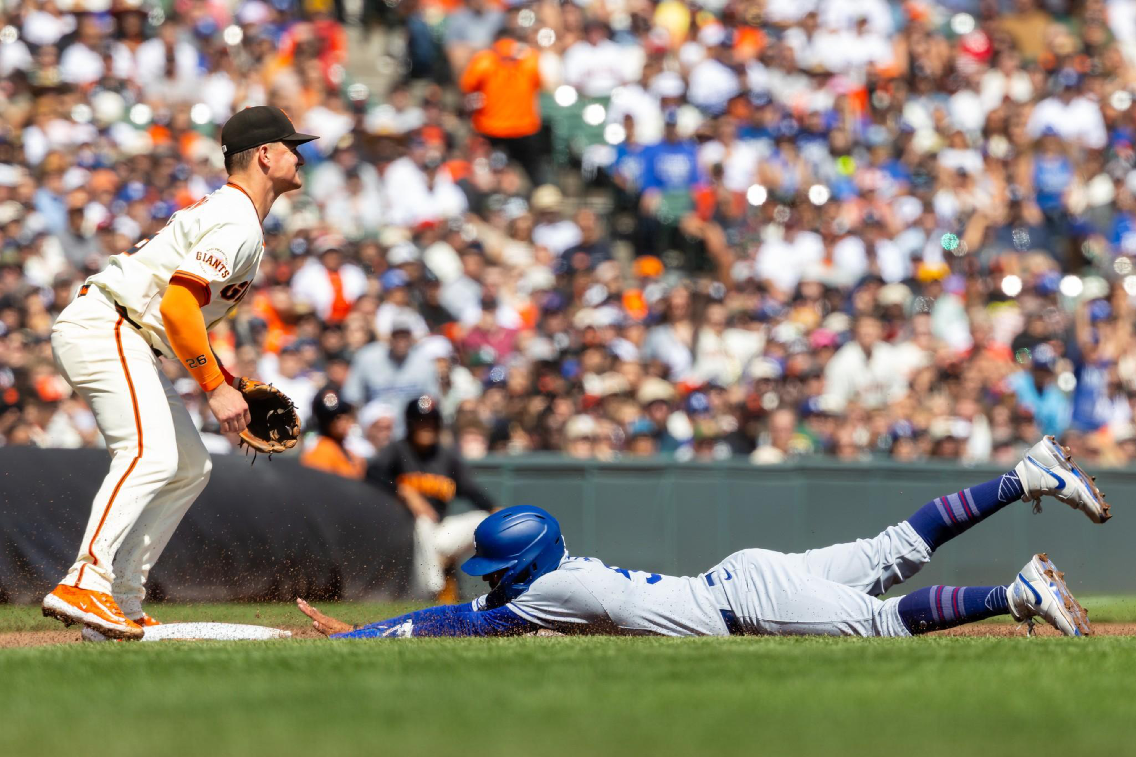 A baseball player in white and blue slides headfirst into a base while a player in white and orange stands ready to catch the ball near the base.