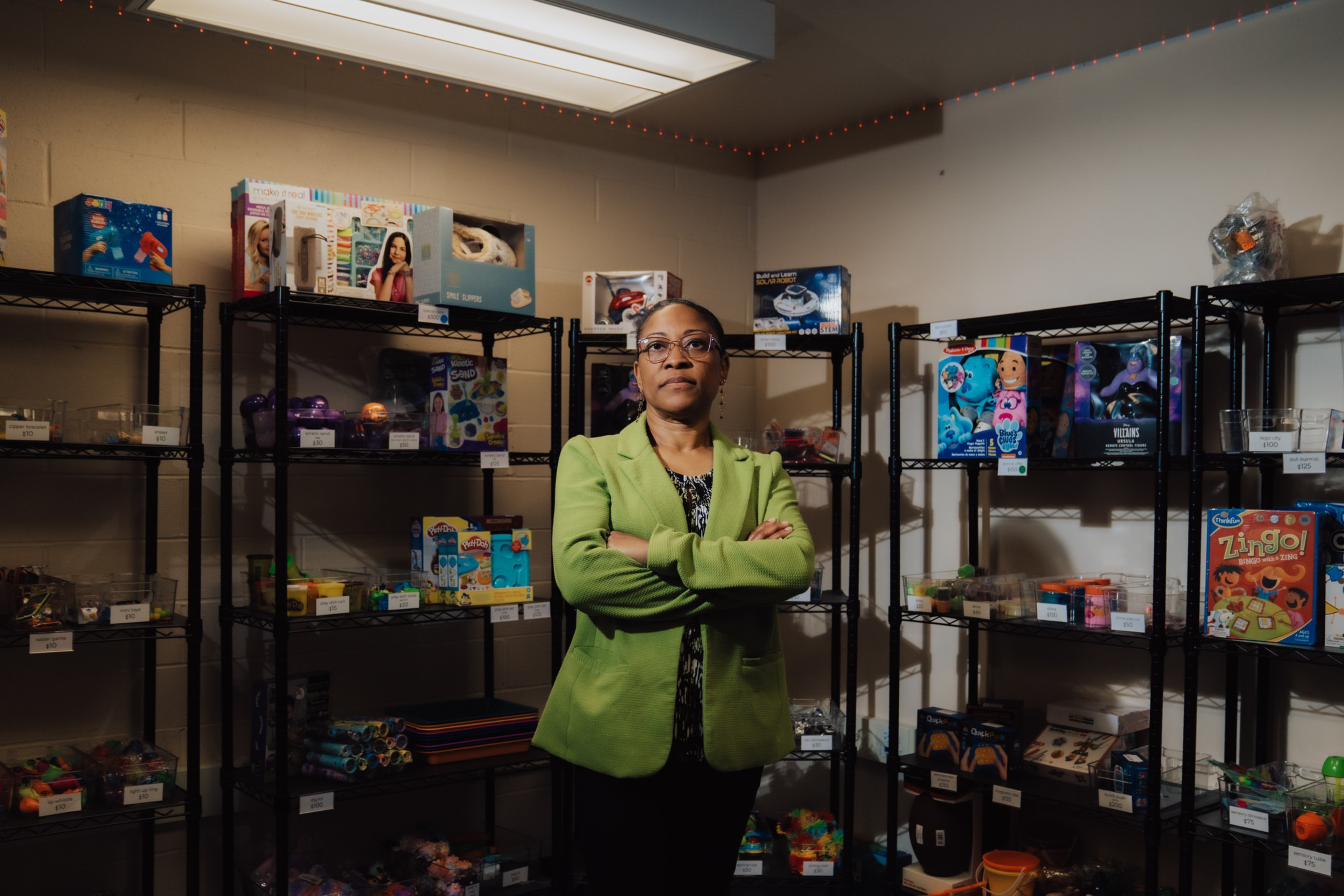 A woman in a green blazer stands with crossed arms in a room filled with black metal shelves holding various toys, games, and craft supplies.