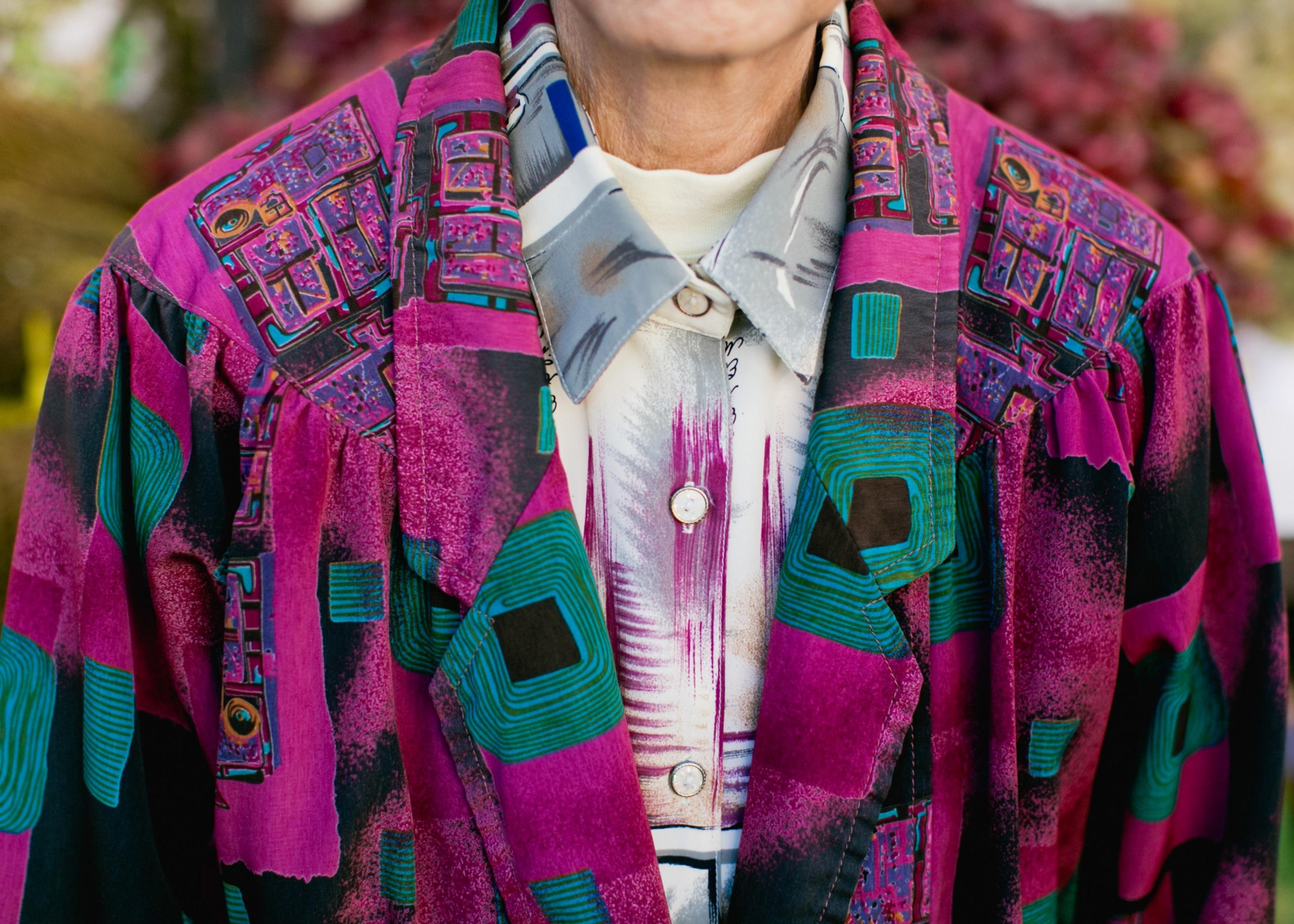 A person wears a vibrant jacket with purple, teal, and black abstract patterns over a white shirt with a gray, purple, and black design.