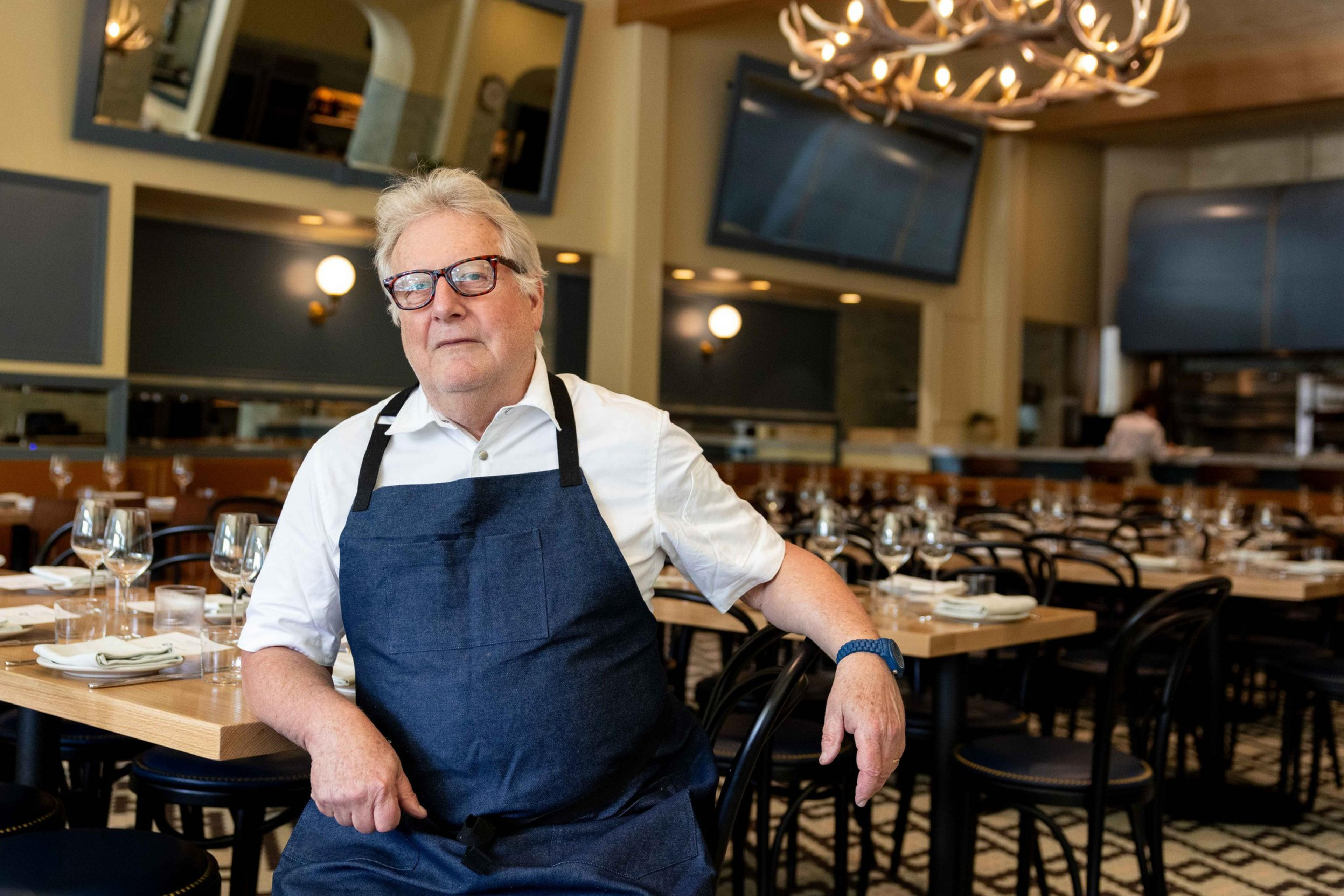 A person wearing glasses and a blue apron stands beside a table in a restaurant with elegant decor, including chandeliers and neatly set tables.