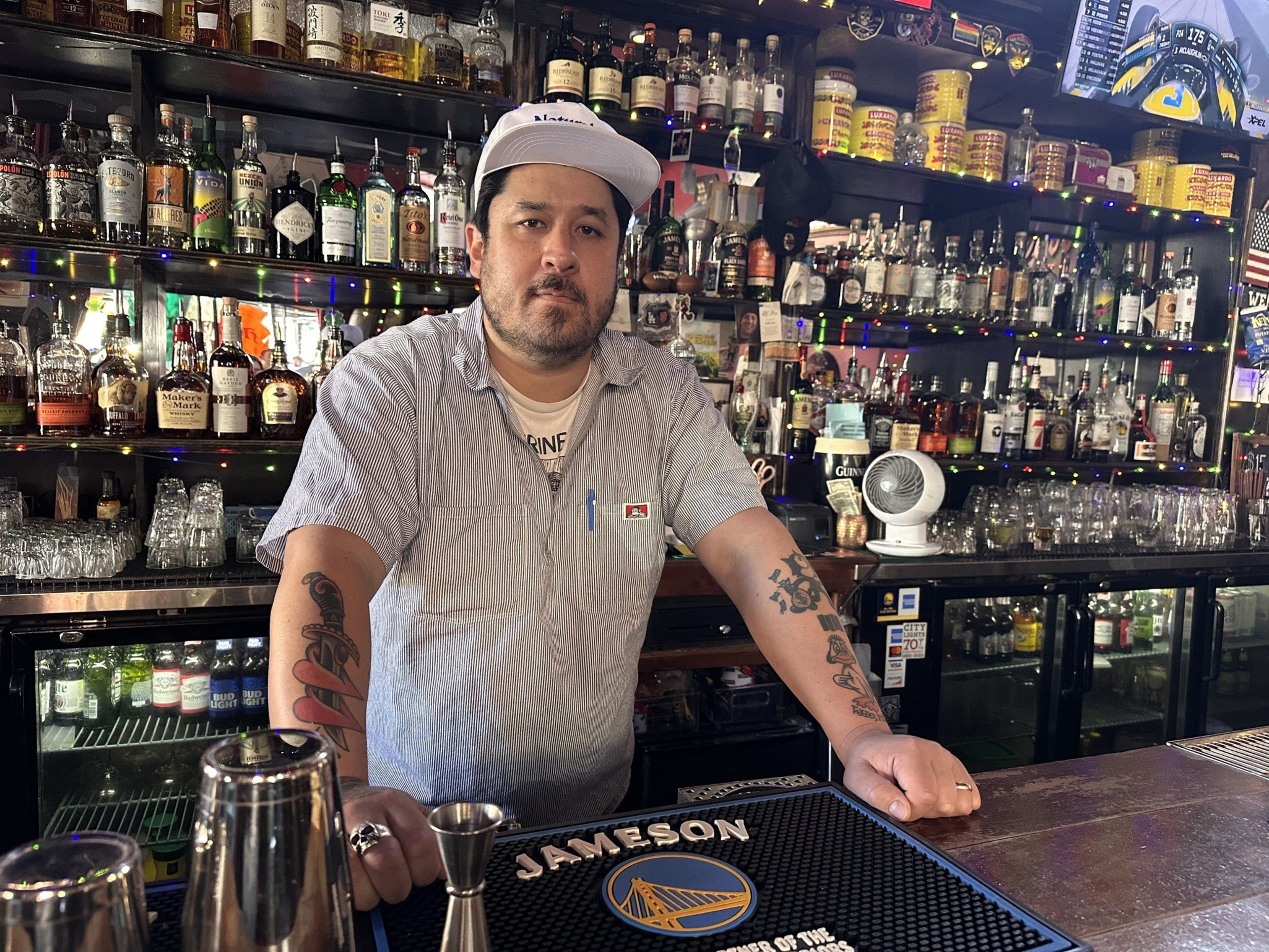 A person stands behind a bar, surrounded by shelves filled with various bottles. They wear a cap and a striped shirt, and have tattooed arms resting on the counter.