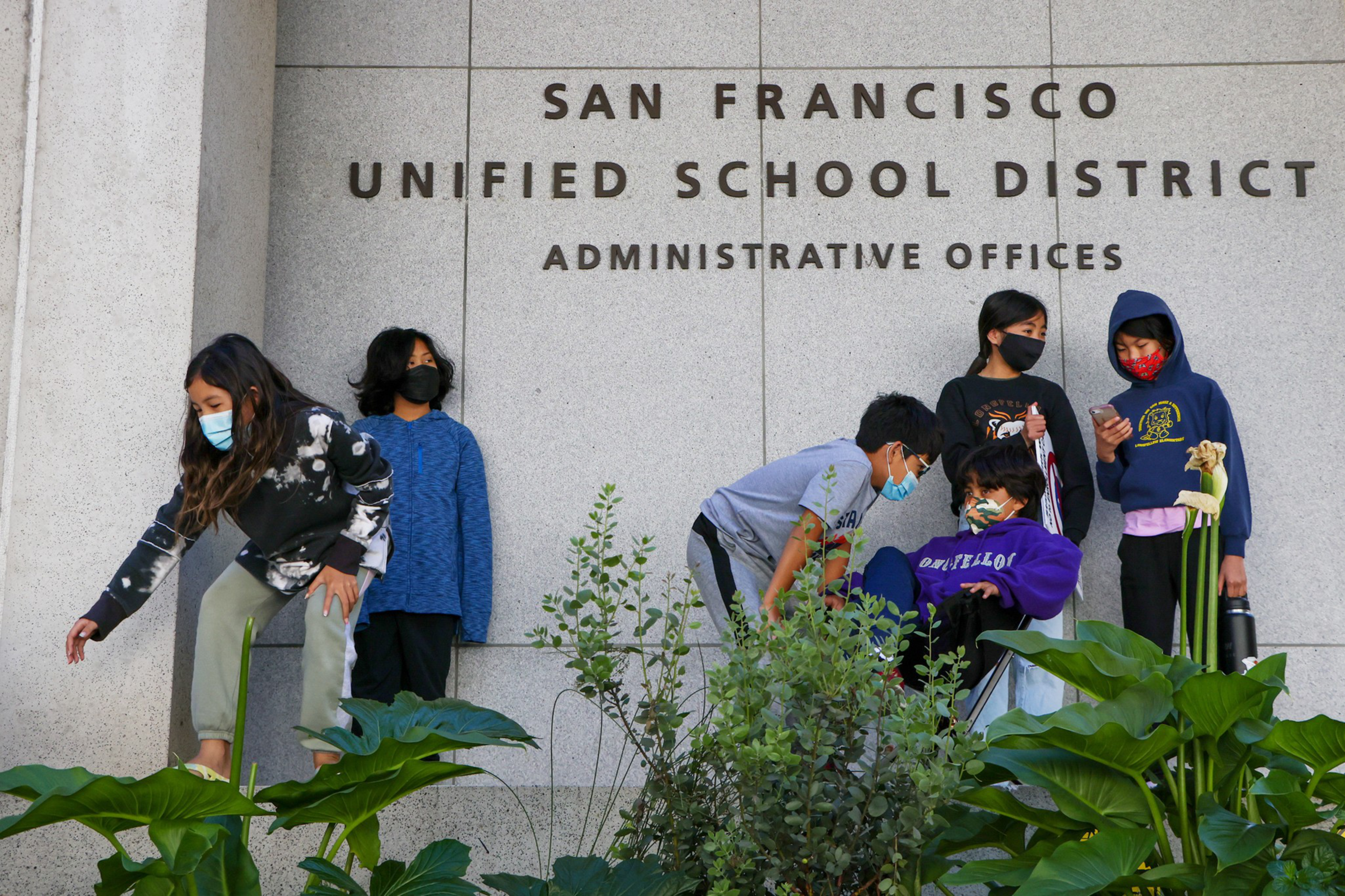Six children wearing face masks stand or sit near a wall with the words "San Francisco Unified School District Administrative Offices." Green plants are in the foreground.