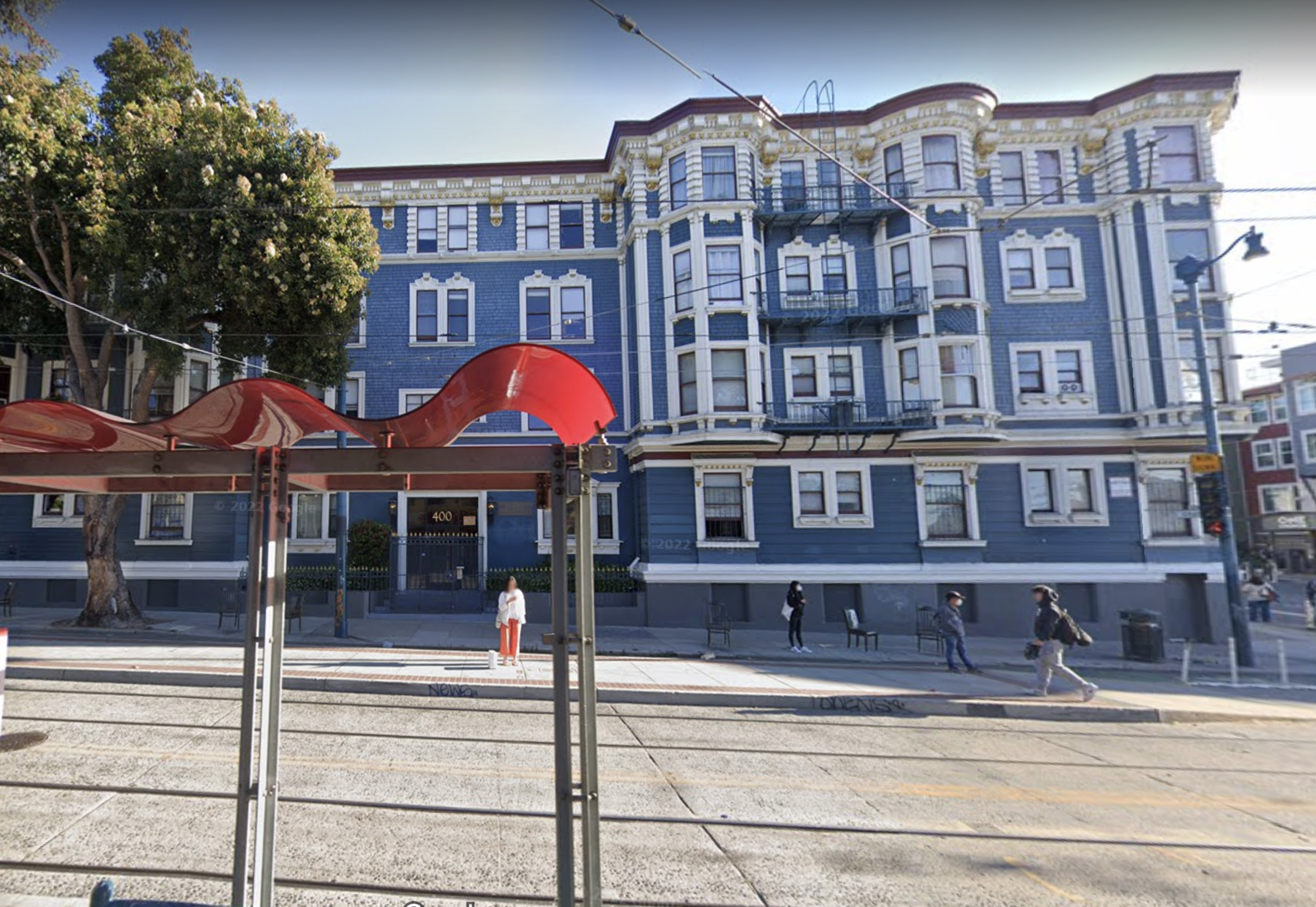 A blue and white multi-story building with ornate trim stands behind a streetcar stop featuring a red wavy roof, with five people waiting nearby.