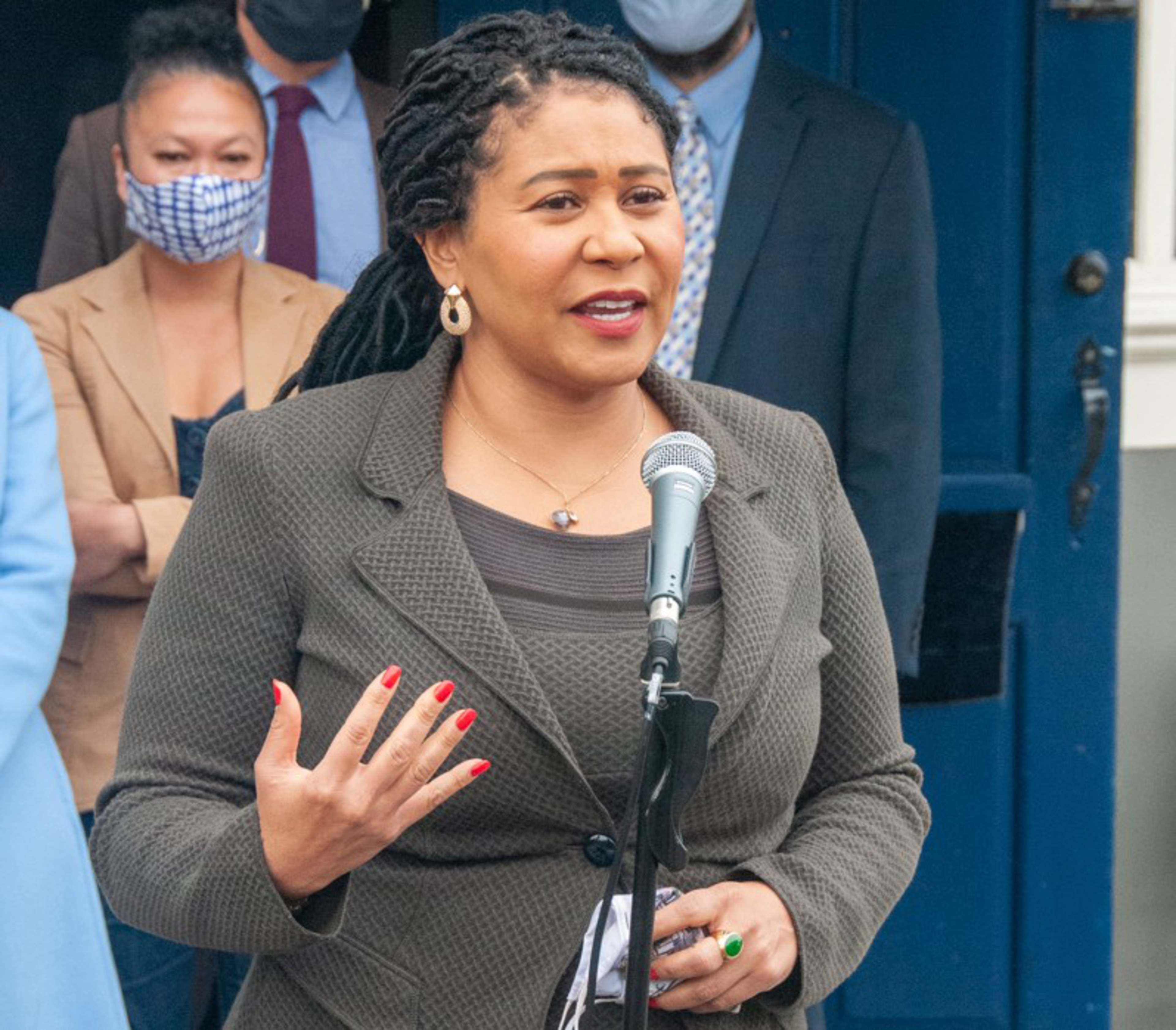 A woman with braided hair speaks at a microphone wearing a gray blazer, while people stand behind her, some wearing face masks.