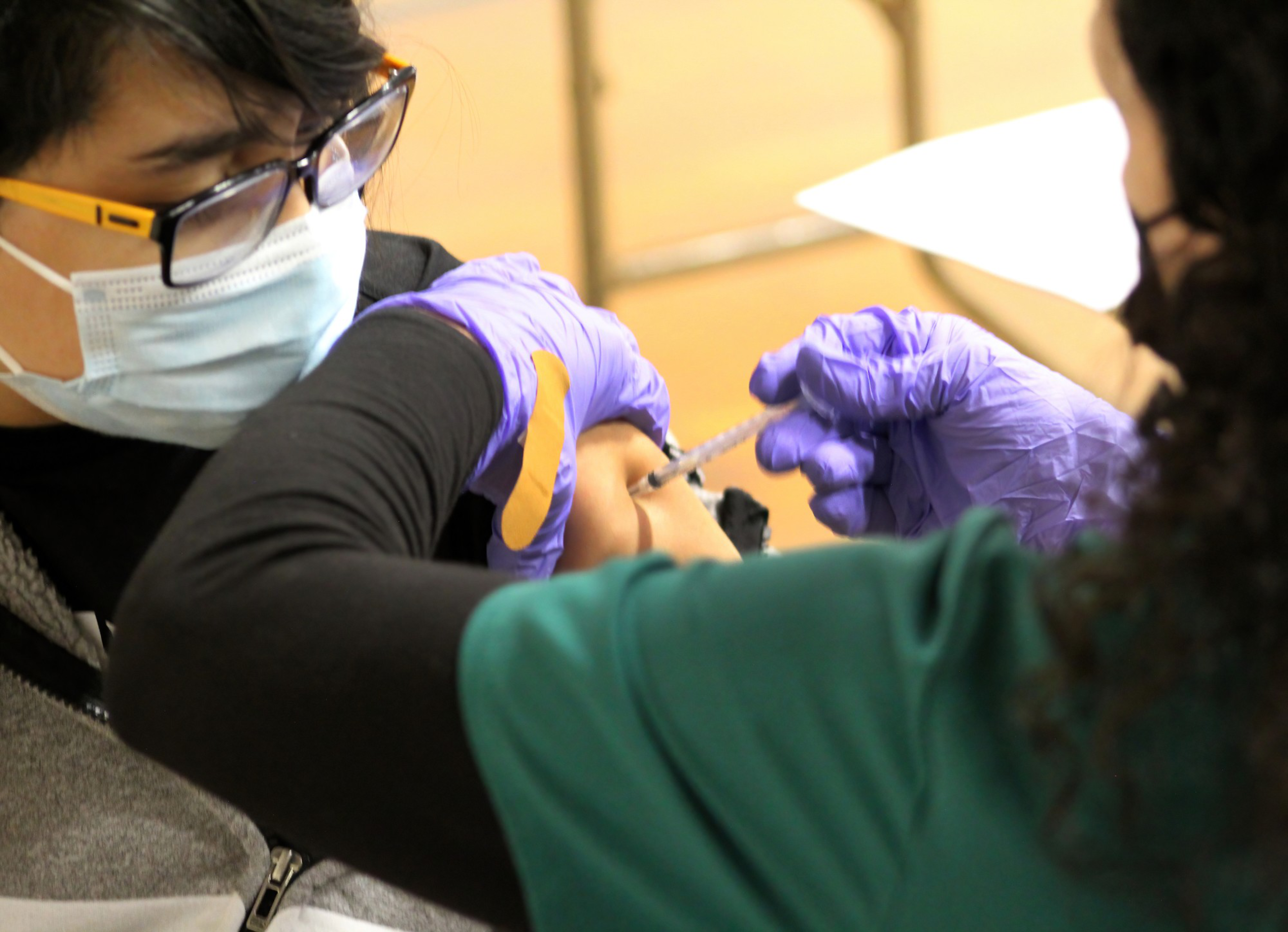 A person wearing glasses and a mask receives a vaccine injection in the arm from a gloved healthcare worker.