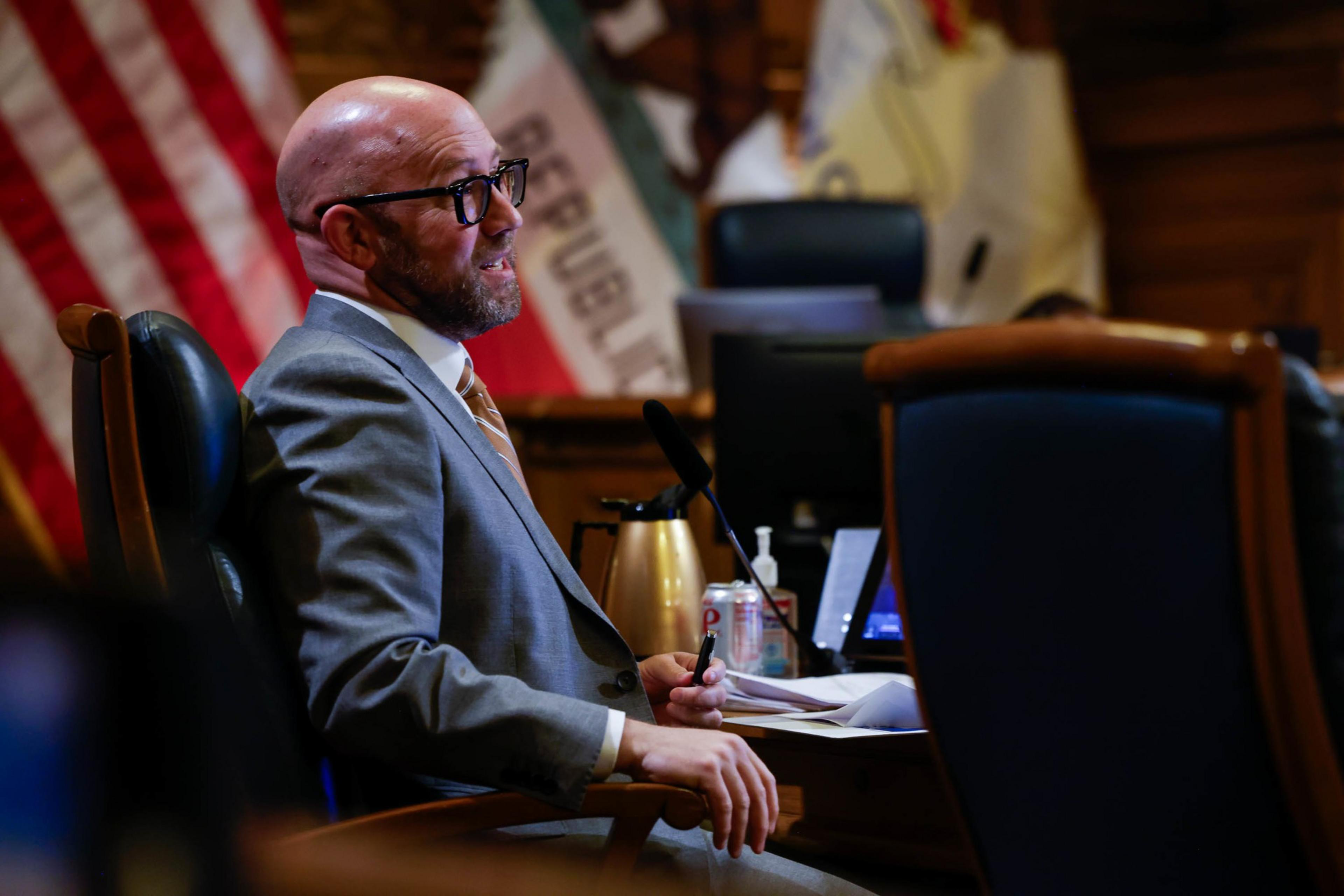 A bald man with glasses, wearing a suit, sits by a flag in a wood-paneled room, with a microphone and papers in front of him.