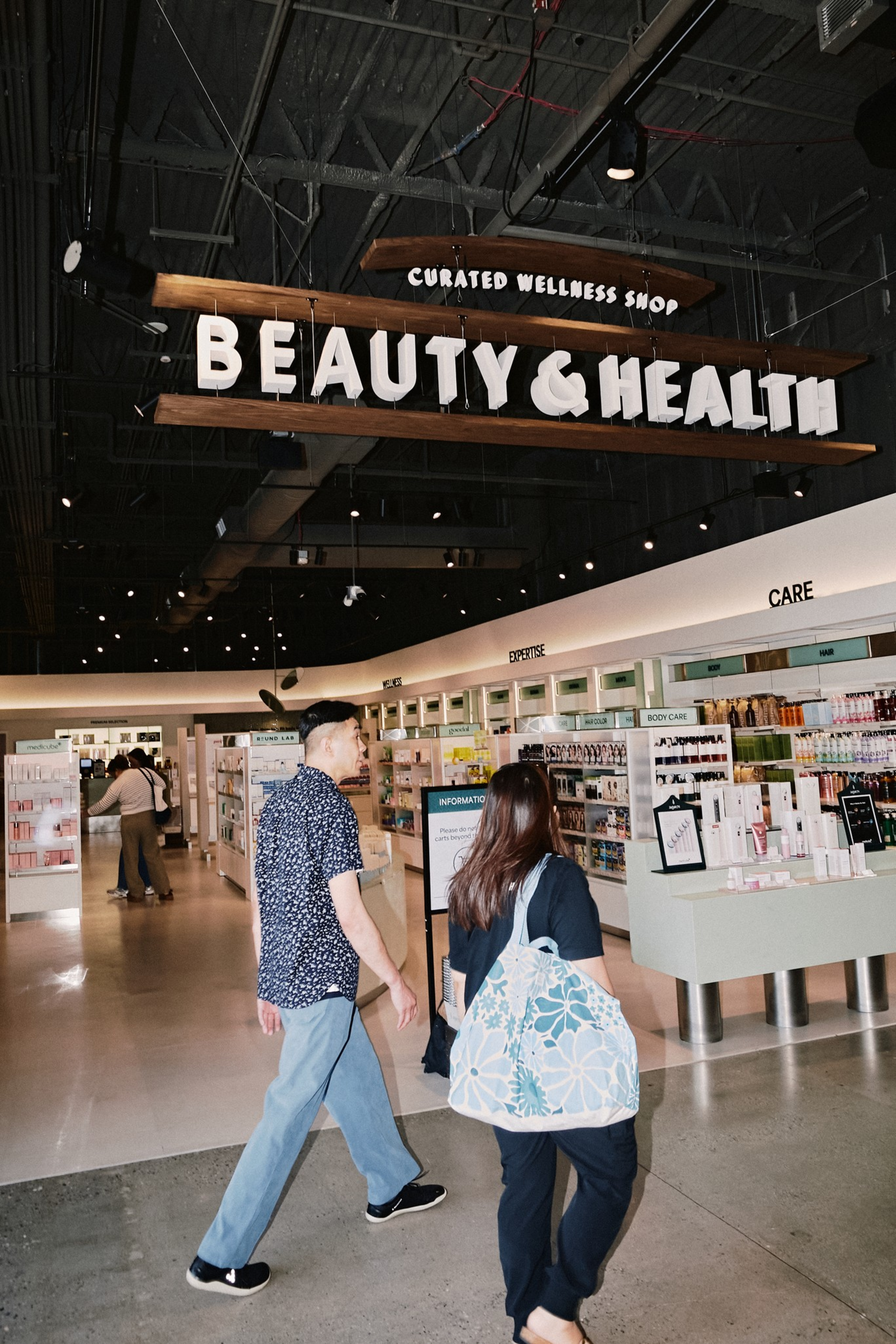Two people walk inside a well-lit store under a large hanging sign that reads "Curated Wellness Shop BEAUTY & HEALTH." Shelves of products line the walls.