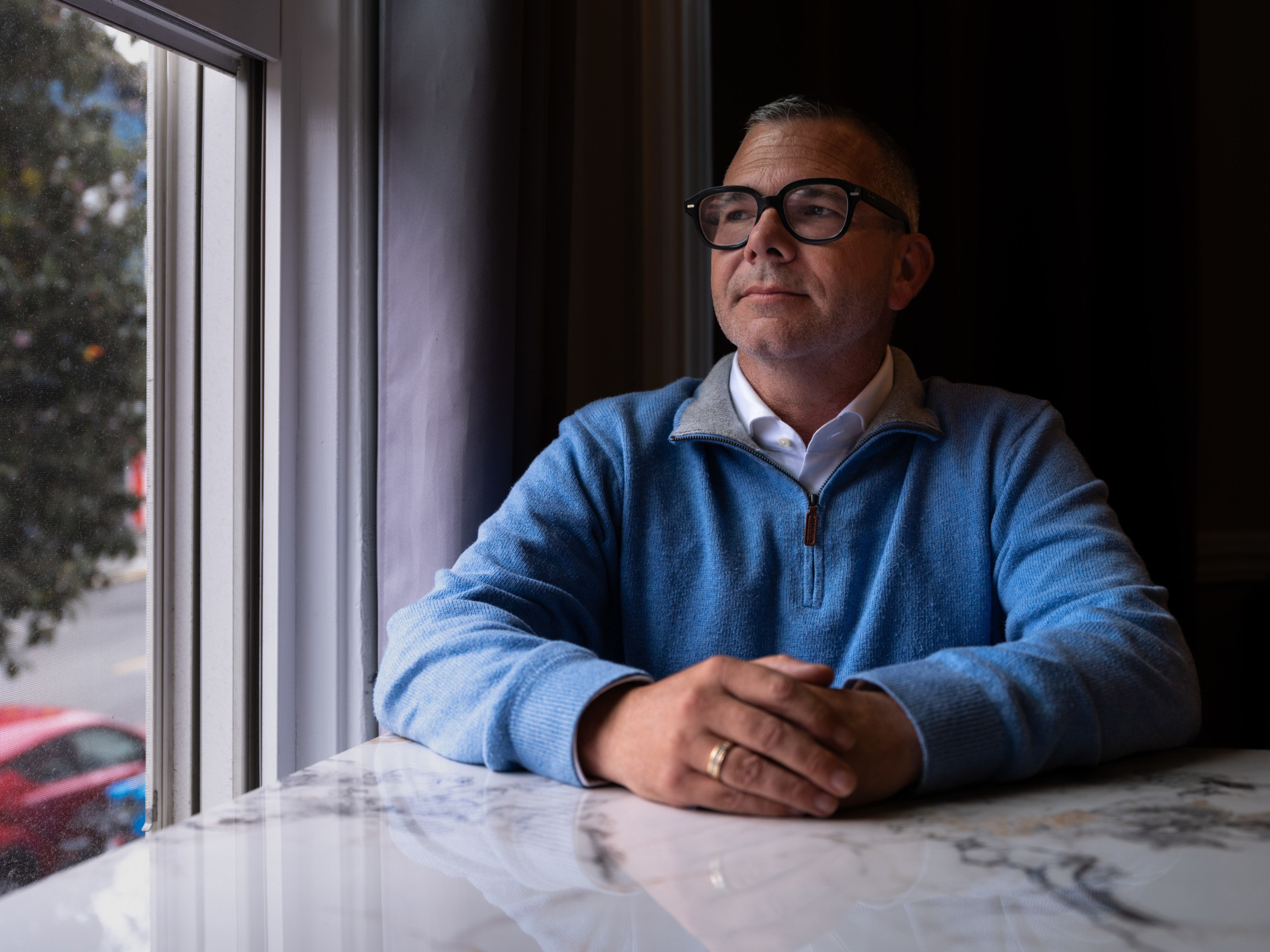 A man in glasses and a blue sweater sits at a marble table, looking thoughtfully out a nearby window with his hands clasped.