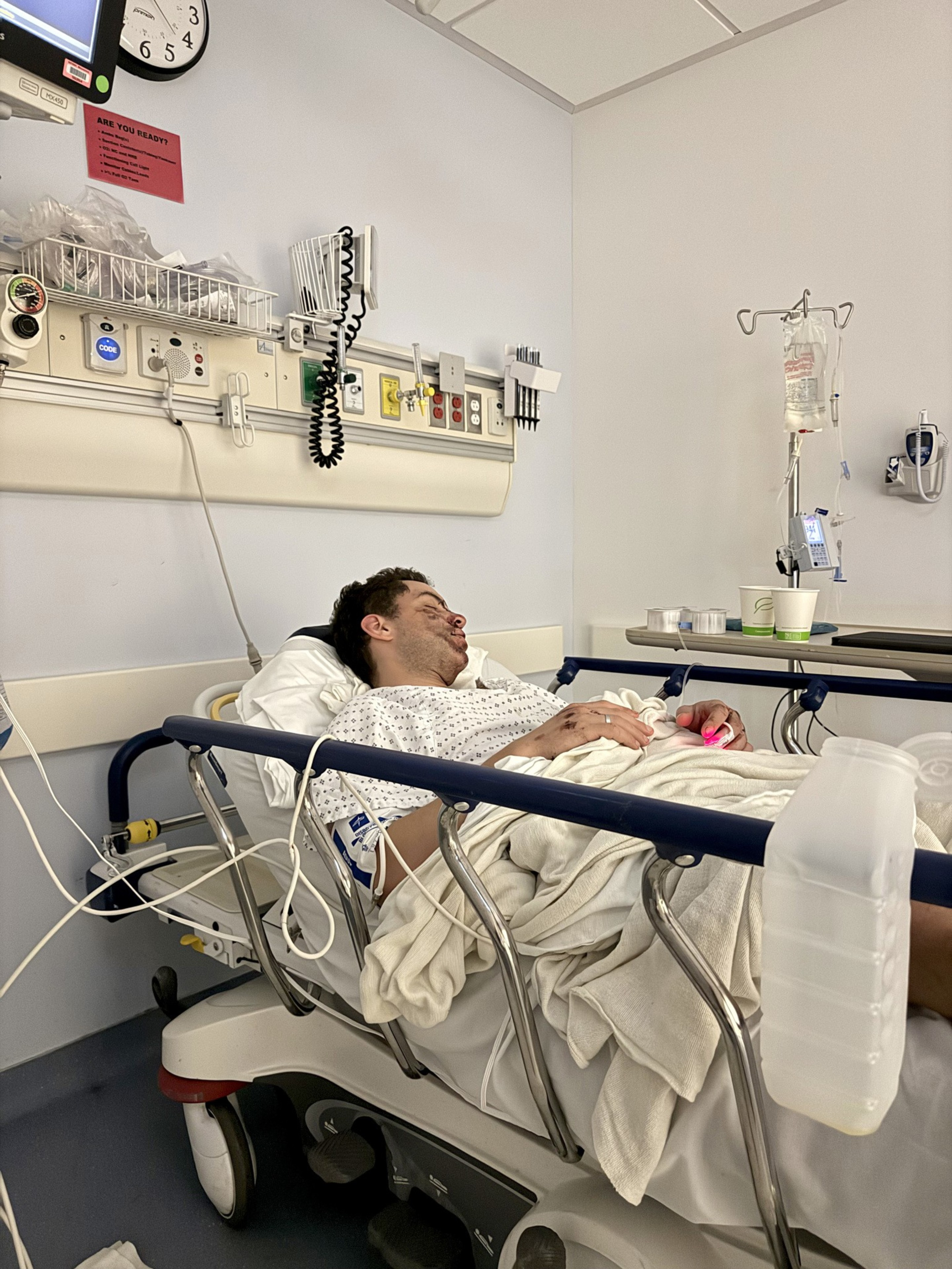 A man lies on a hospital stretcher with an IV drip, wearing a hospital gown, covered by a blanket, resting in a clinical room.