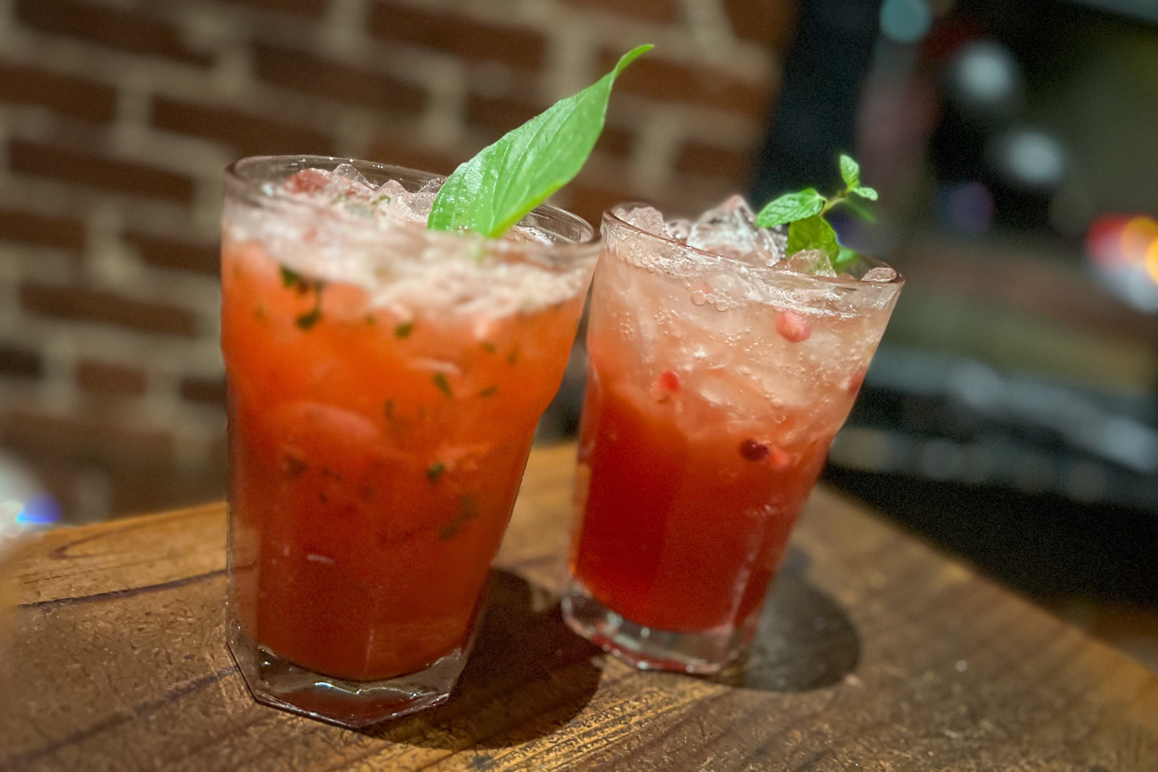 Two red, iced cocktails garnished with green mint leaves sit side by side on a wooden surface, against a blurred brick wall background.