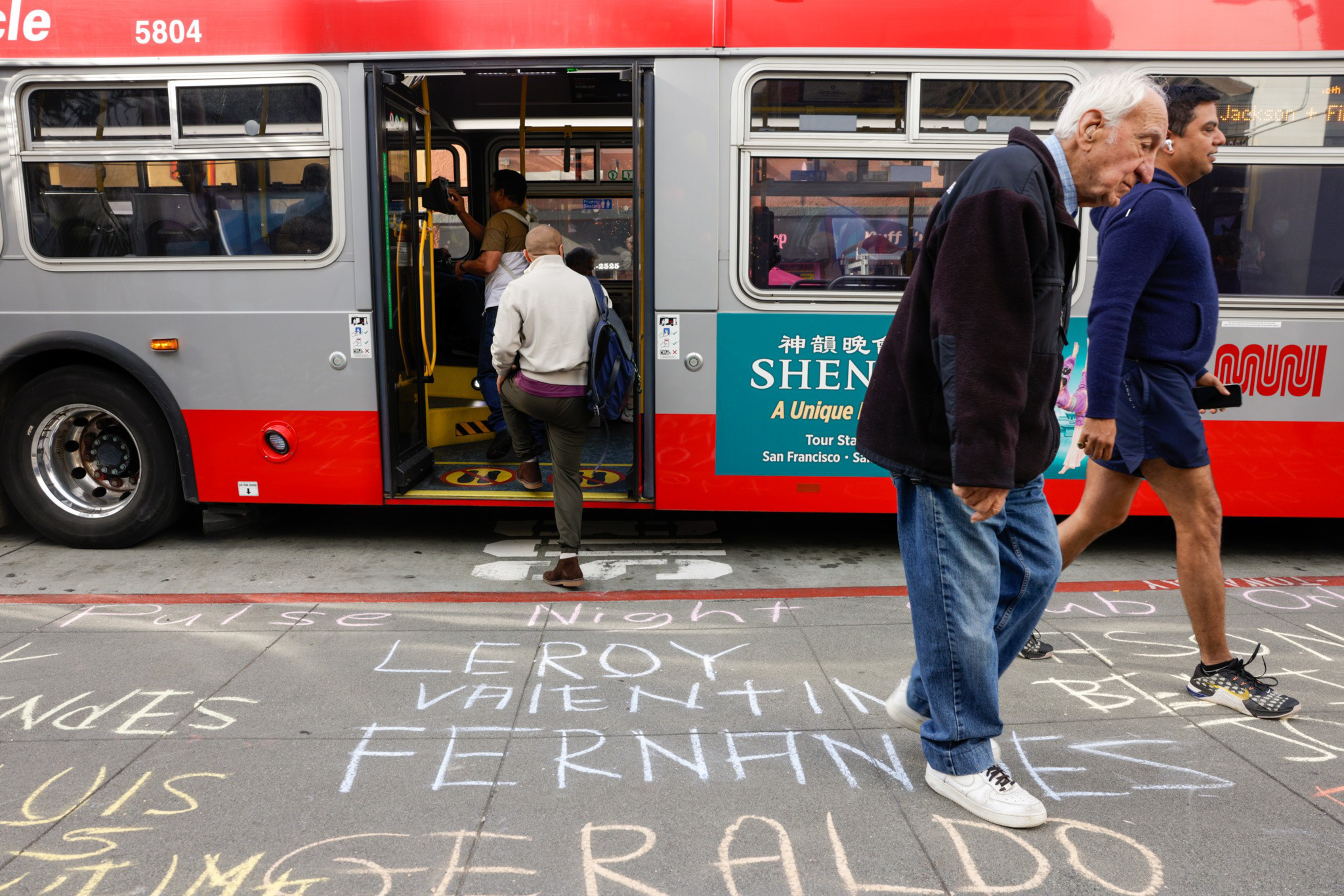 A bus stop where people on gettting on and off while the sidewalk is covered by peoples names with chalk that have died because of AIDS.