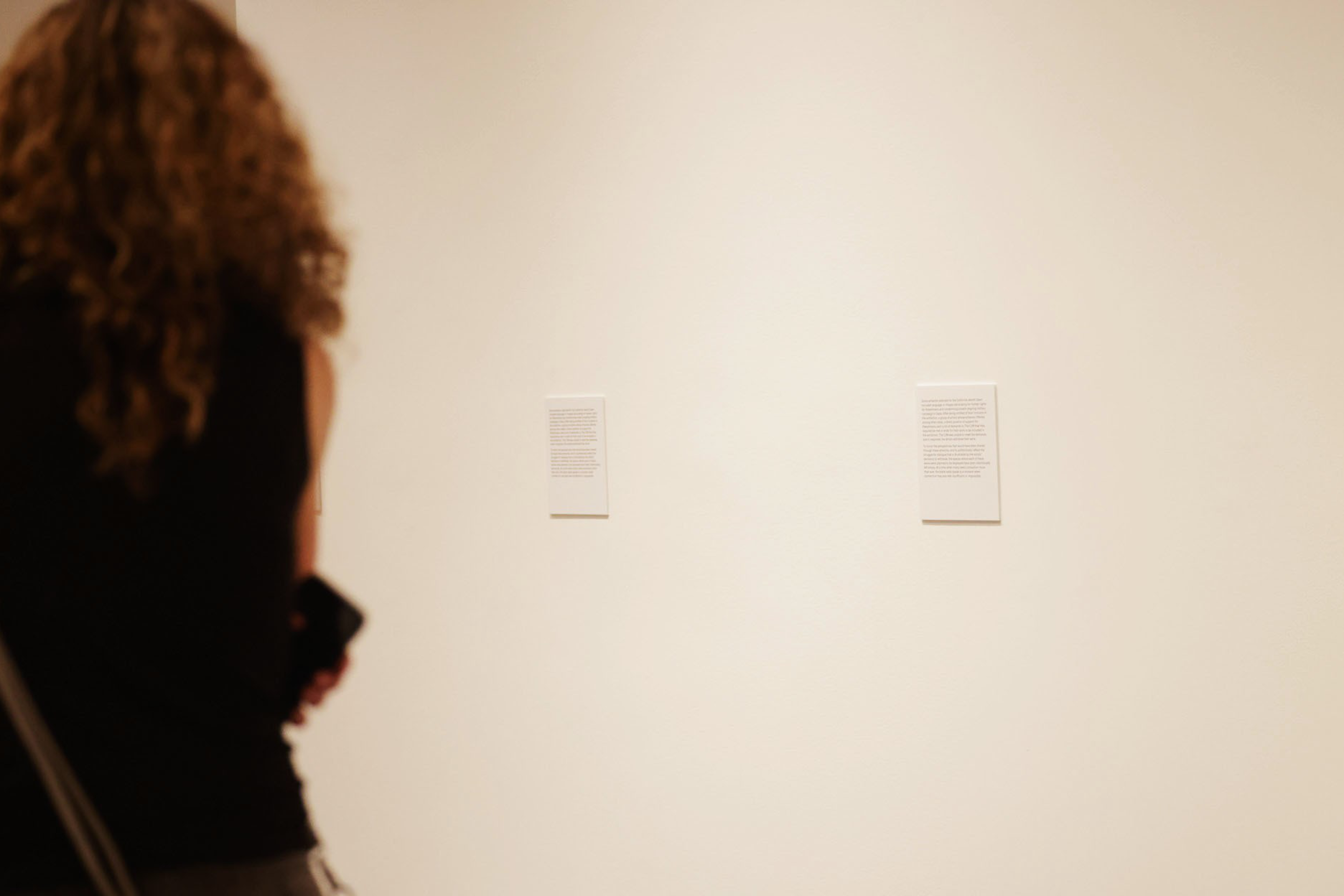 A person with curly hair, dressed in a sleeveless top, stands in front of a beige wall reading two small text panels.
