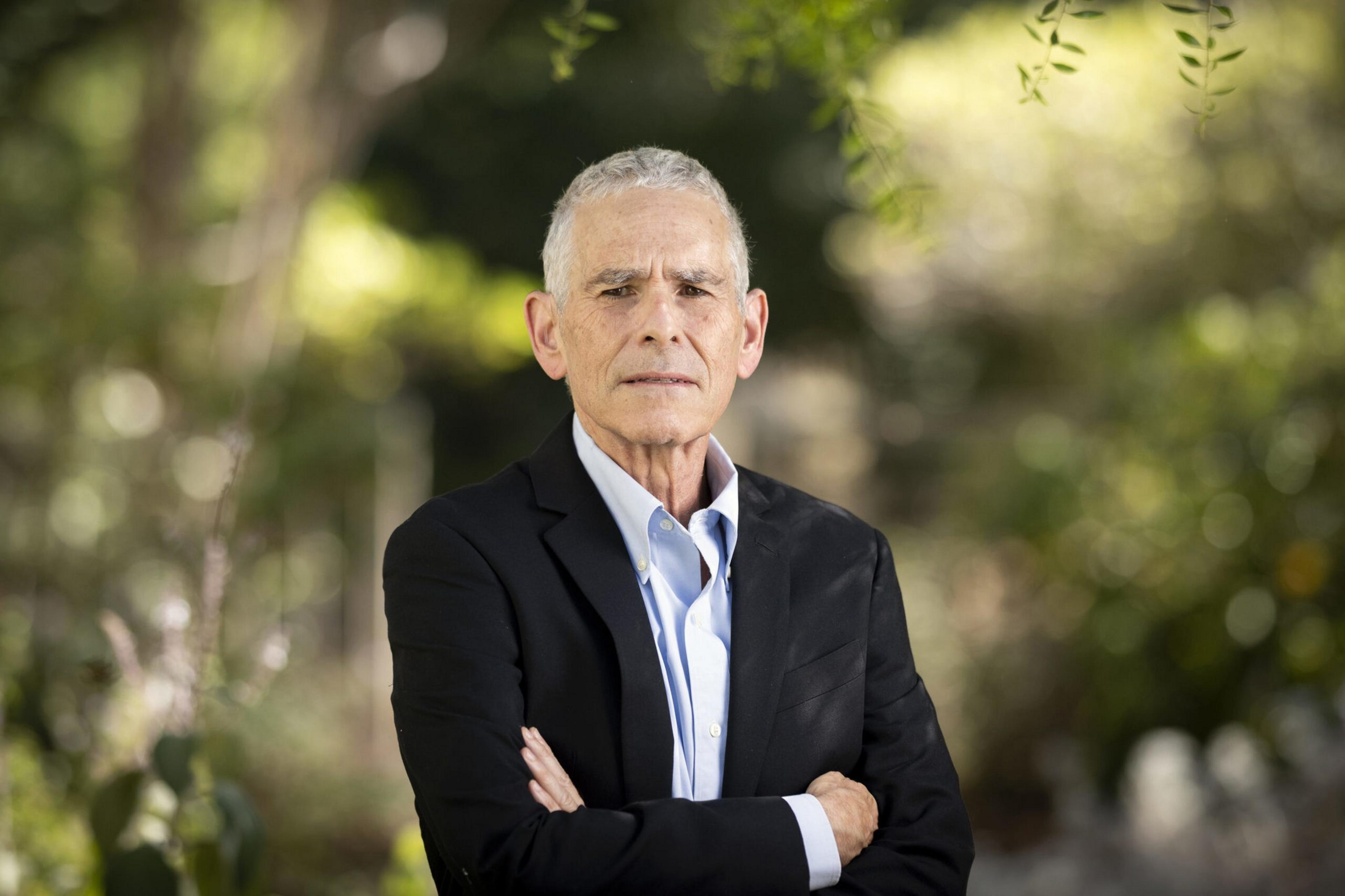 An older man with short gray hair stands outdoors wearing a black blazer and light blue shirt, arms crossed, with a serious expression.