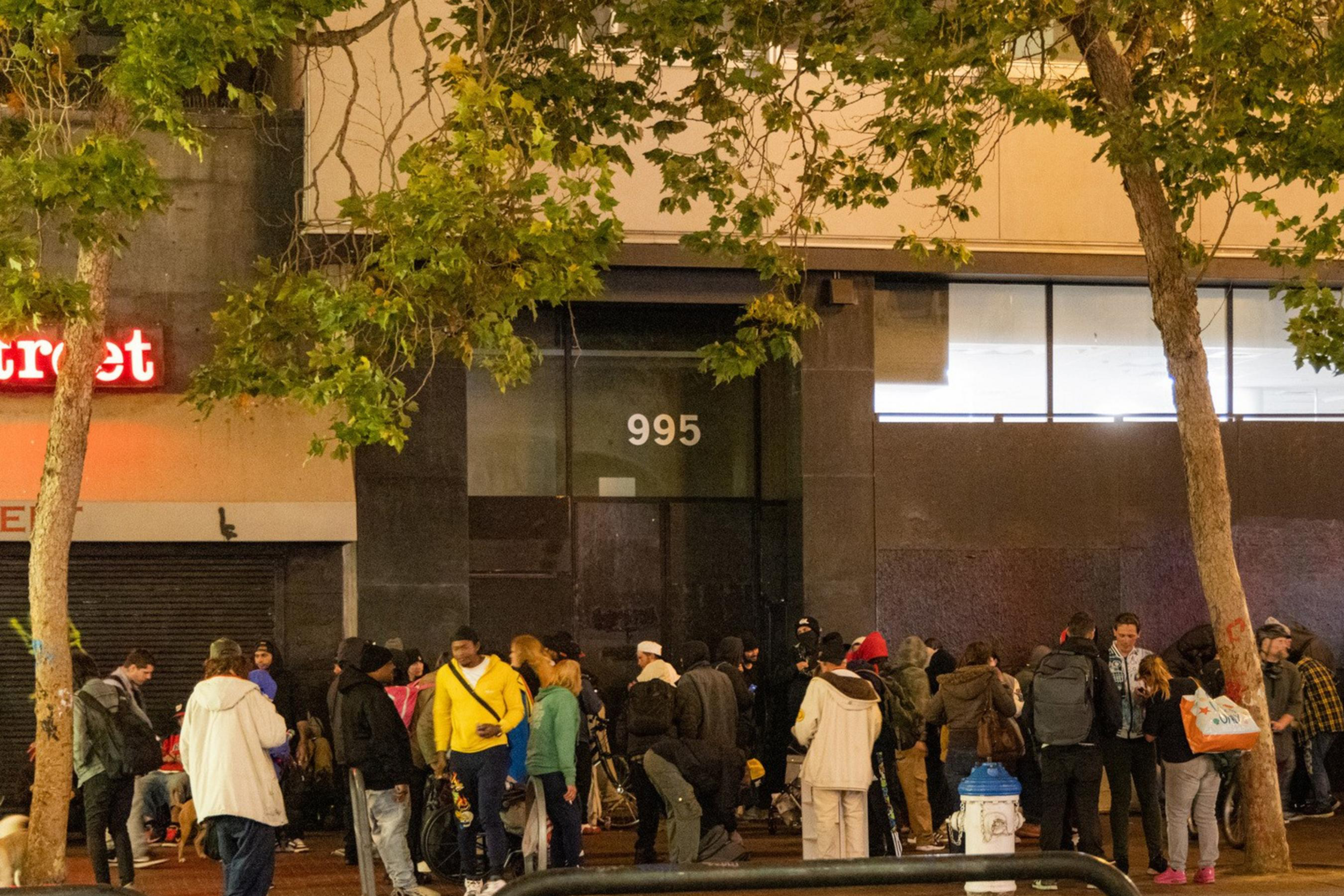 A large crowd of people is gathered on a sidewalk at night near a building with the number 995 above the entrance. Many are wearing coats and hats, and a few trees line the street.