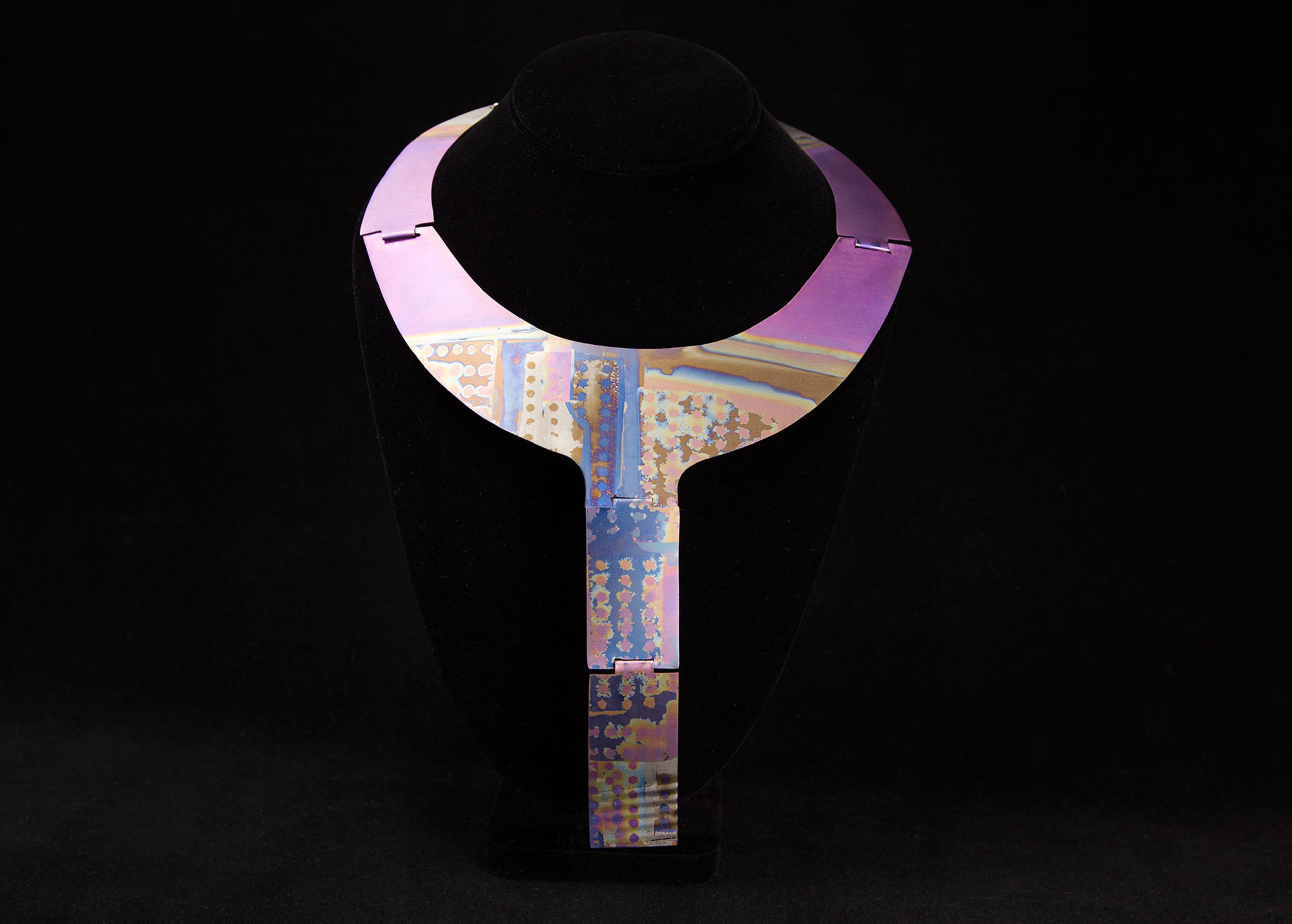 A metallic necklace with an iridescent finish featuring purple, blue, and gold hues is displayed on a black velvet bust.