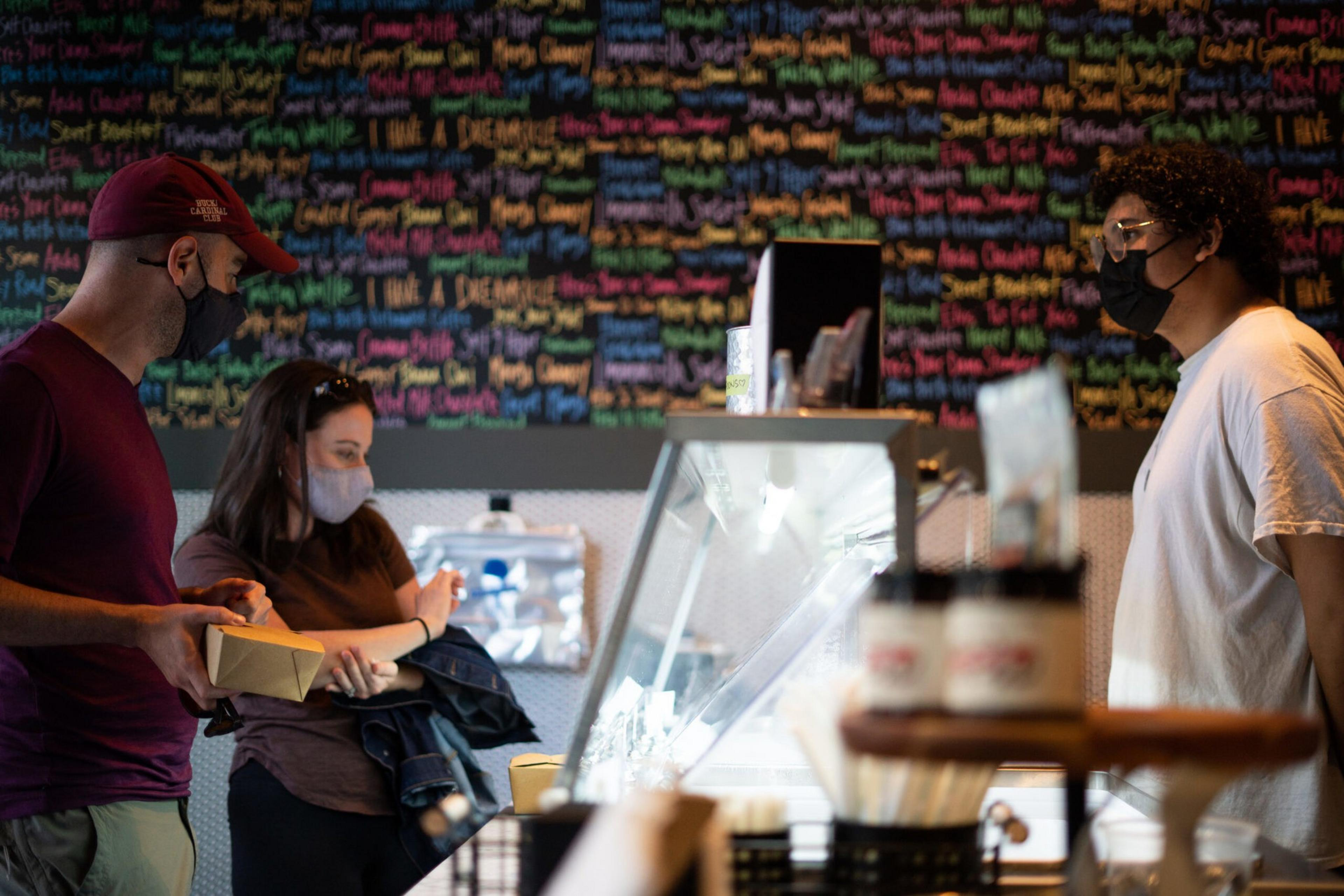 Two masked customers stand at a counter talking to a masked employee in a shop with a colorful menu board behind them.